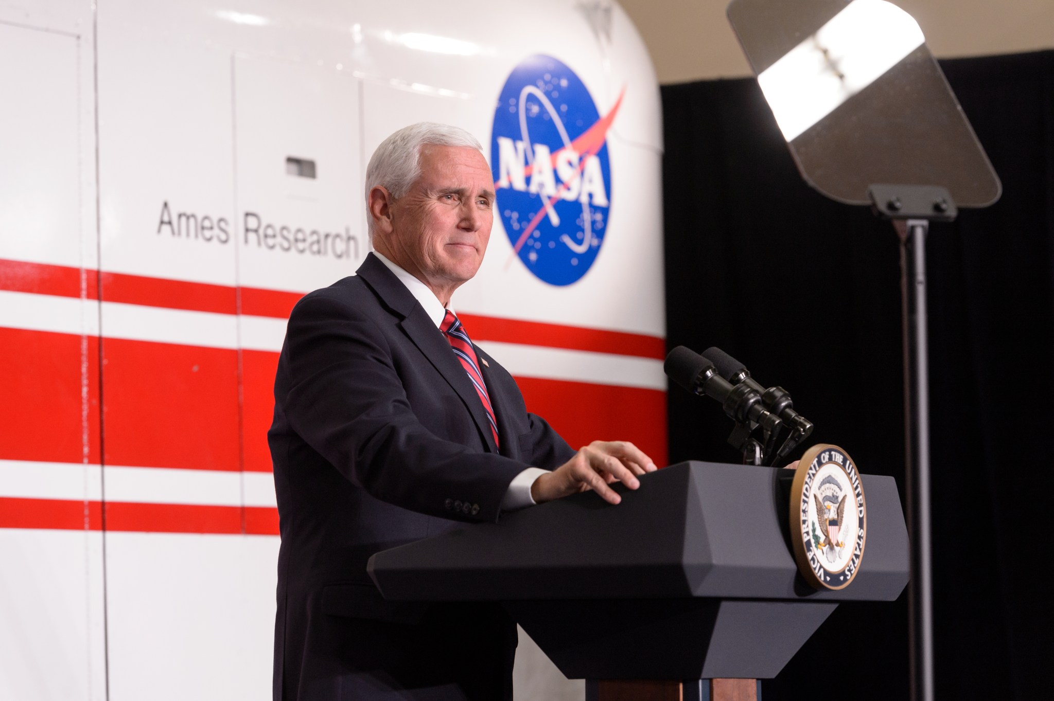 Vice President Mike Pence stands at a podium in front of a white vehicle with red stripe and a NASA logo