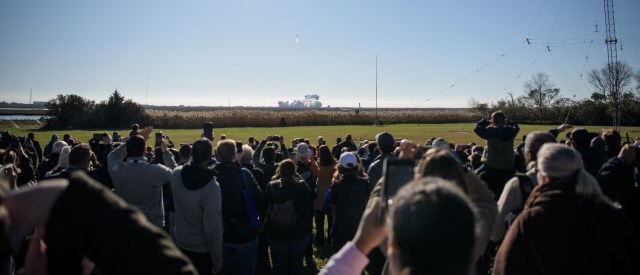 A large group of people, some holding up cellphones, watches the Antares rocket launching in the distance.