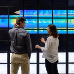 Two people standing front of a wall of screens showing a multicolored image of a rocket body
