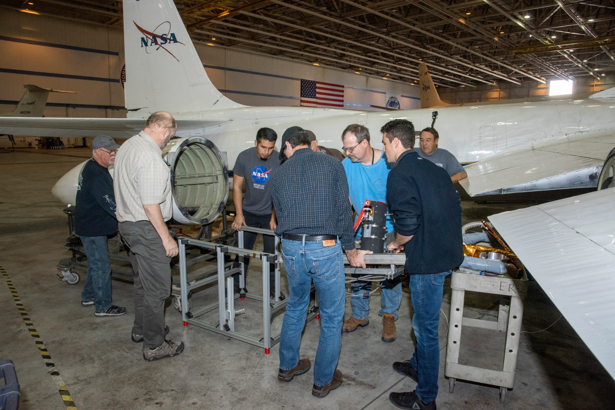 A crew of people loading a mechanical instrument into a casing hanging below one wing of the ER-2 plane.