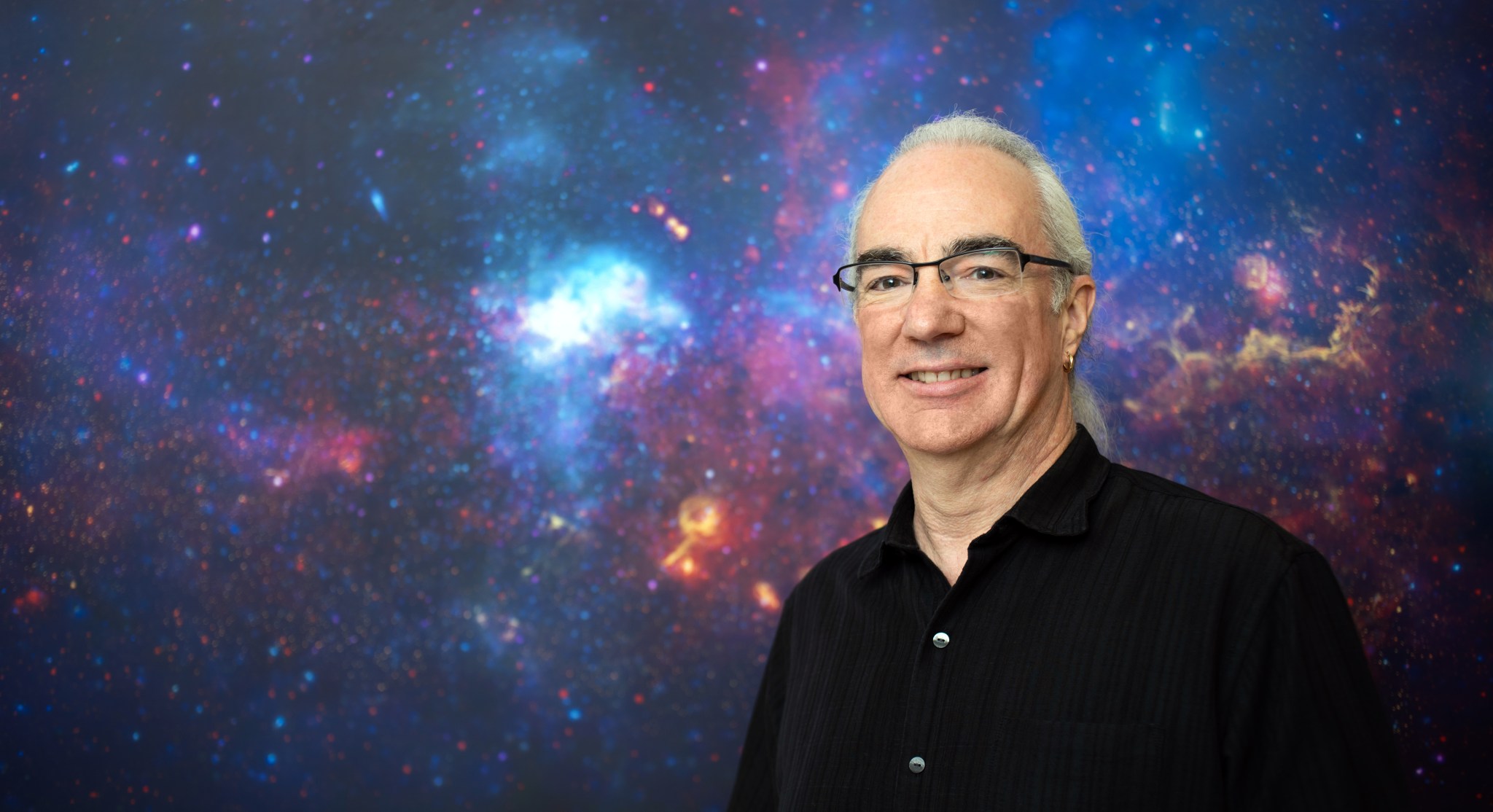 Man with fair skin and long grey hair, pulled back into a ponytail in front of a blue, pink, orange and purple galaxy background. He wears glasses, an earring, and a black collared shirt.