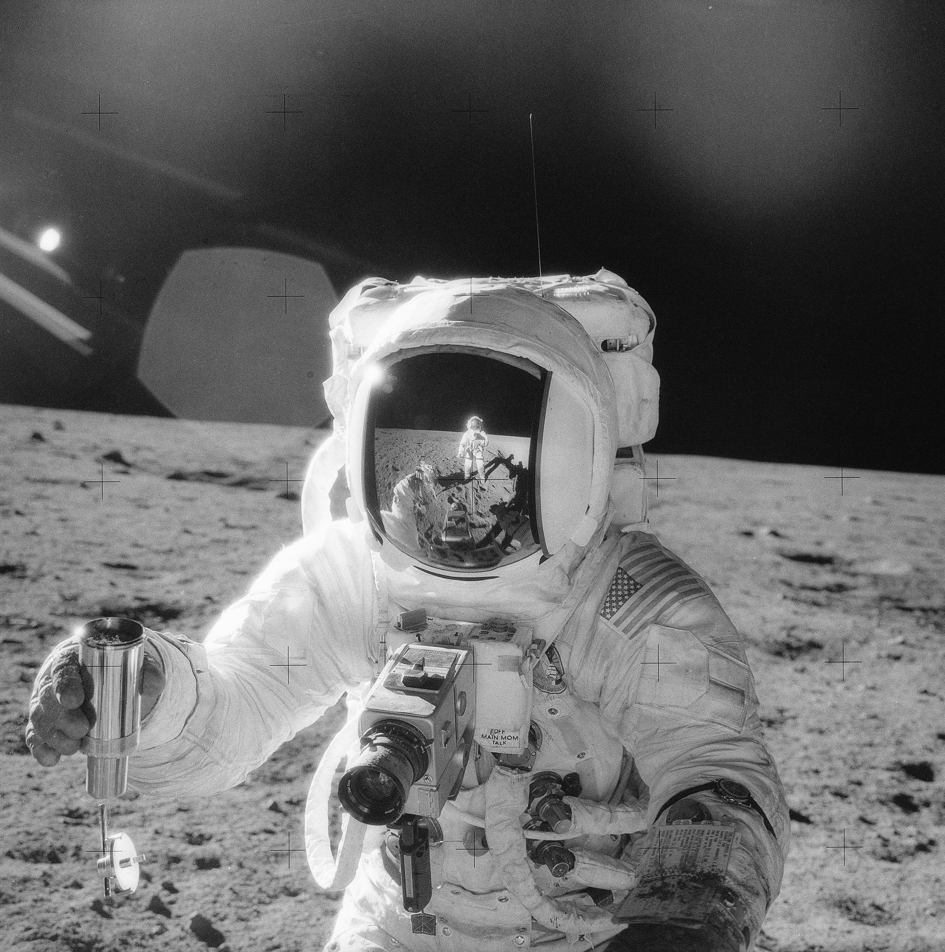One of the Apollo 12 astronauts on the Moon’s surface is holding a container of lunar soil, the other is reflected in helmet. 