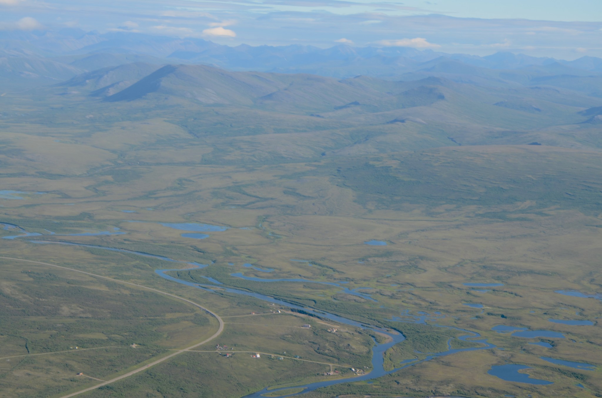 An airborne view of Arctic tundra, with grass and short scrub brush covering most of the ground and thin rivers and small lakes dotting the landscape.