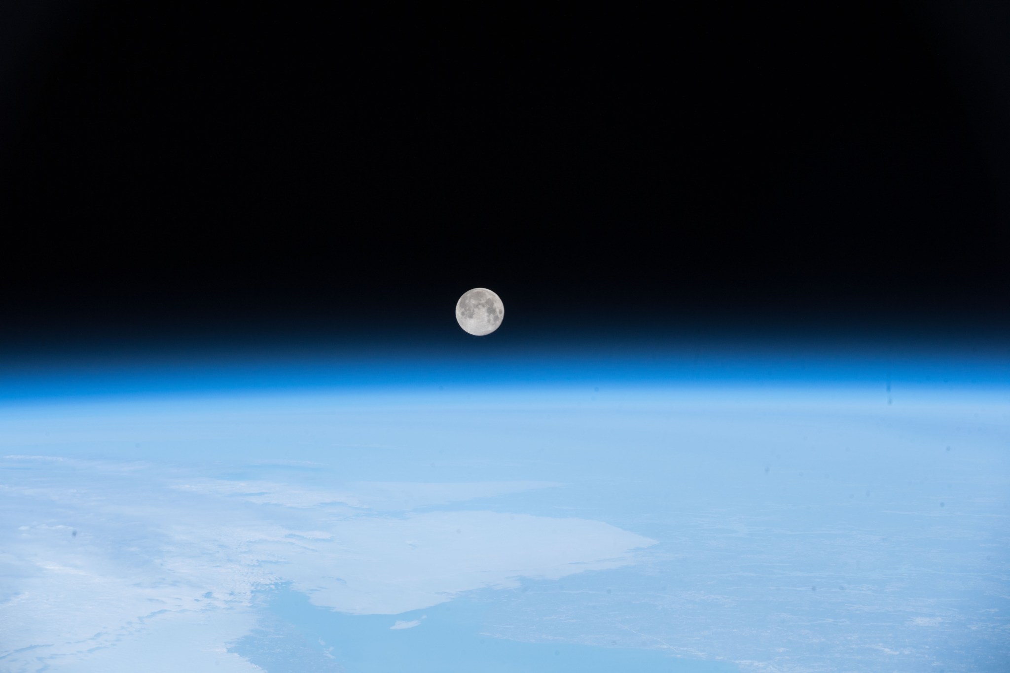 The Moon, a silvery white globe, floats above the diffuse blue atmosphere of Earth, which curves gently across the image, cutting it in half. The top half is the black of space.