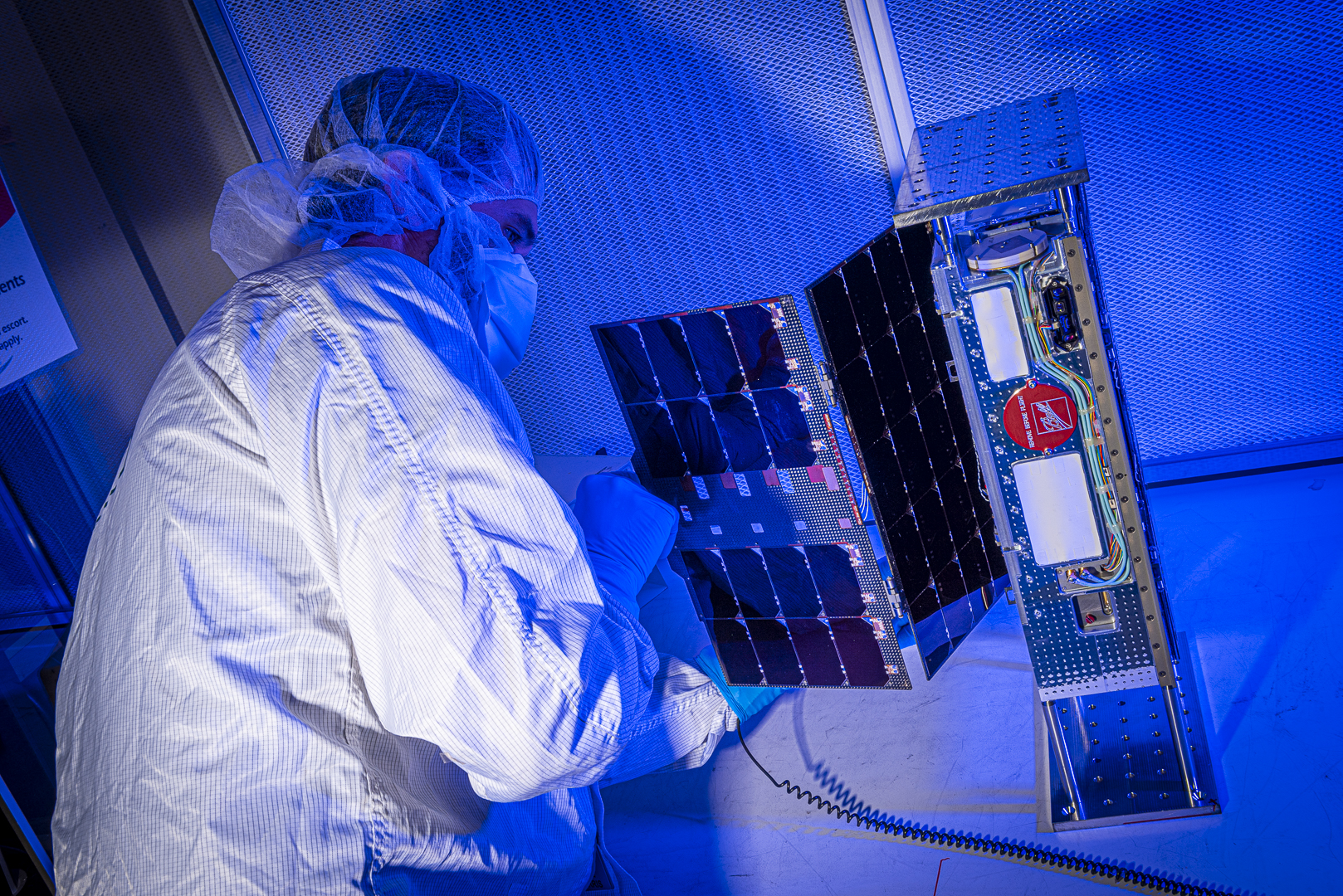 A technician in a white lab coat with a hairnet and mask leans over a CubeSat. The satellite is a thin rectangle, like a computer tower, with electronic components on the near side. Out one longer side, a solar panel is partly unfurled. The tech is inspecting the solar panel. The entire scene is bathed in blue light.
