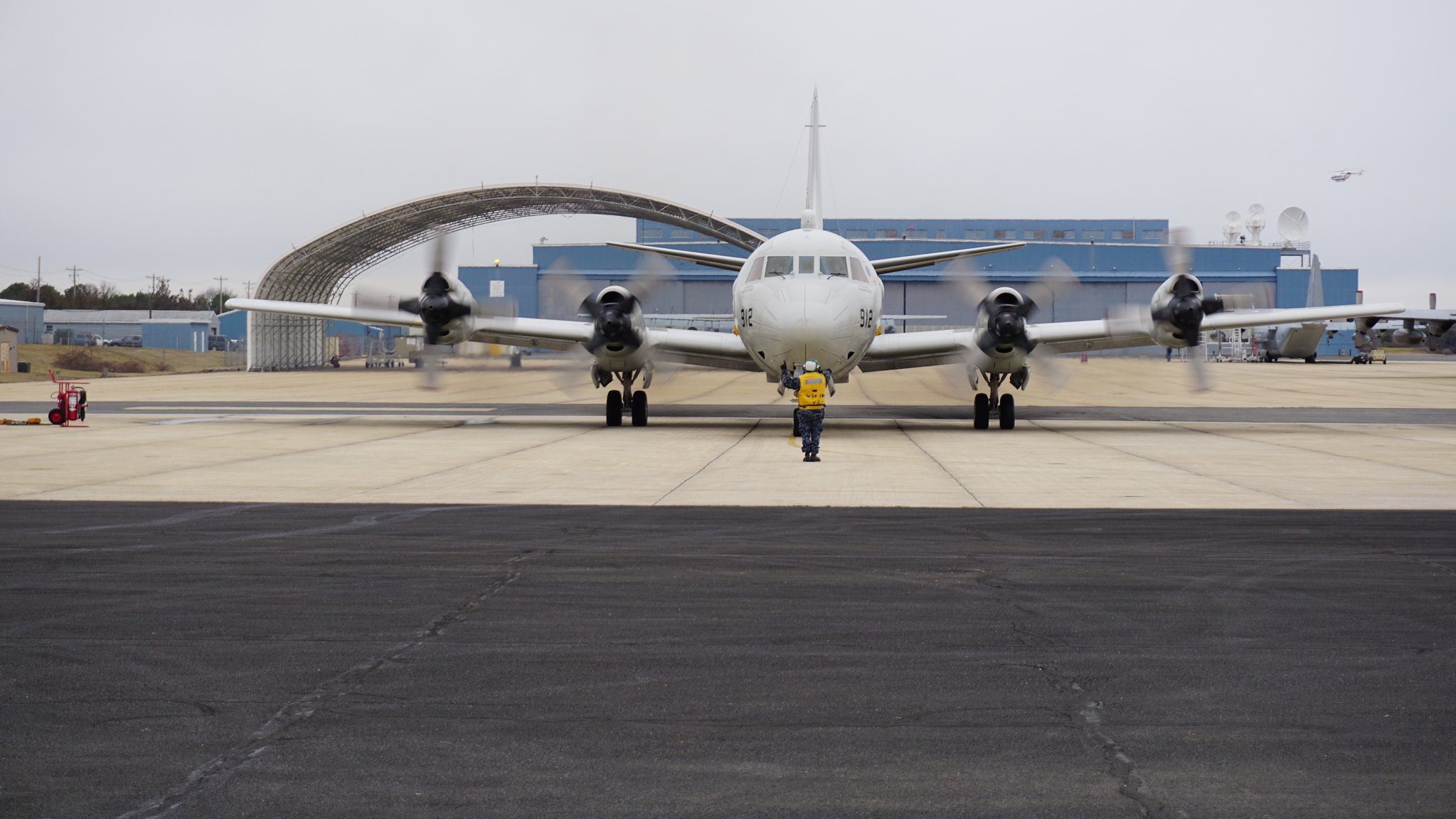 NASA's DC-8 plane on a runway facing the camera against a gray sky. The planes four propellers on its wings are turning. A person stands in front of the nose of the plane.