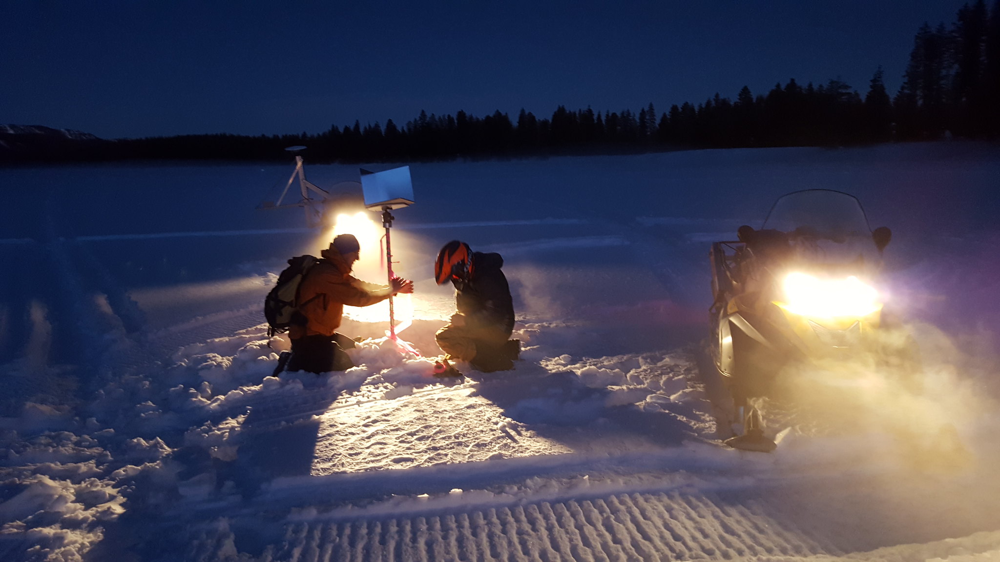 Two people in winter gear kneeling in snow. They are lit by a the headlights of two snowmobiles, one behind the two people and one to the side. 