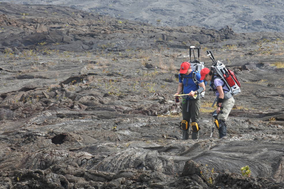 Two people wearing backpacks and other gear in the middle of a barren landscape