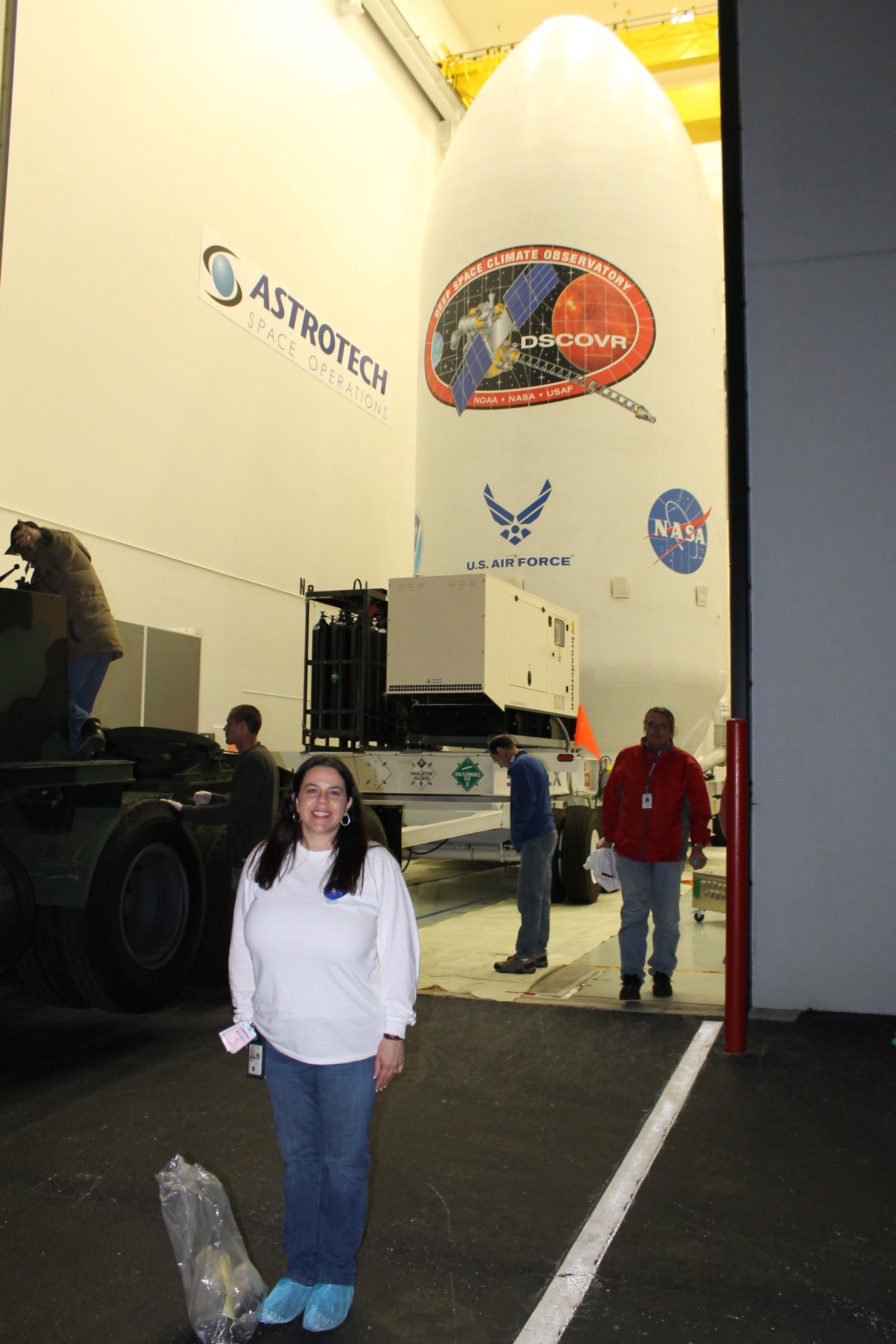 Woman with light tan skin and shoulder length black hair wears  a white long sleeve t shirt, jeans, blue sterile shoe covers and silver hoop earrings. She stands in front of a large rocket with a satellite logo that says "DSCOVR" "Deep Space Climate Observatory" "NOAA NASA USAF" The rocket is on a vehicle to move it and there are four men in the background. 
