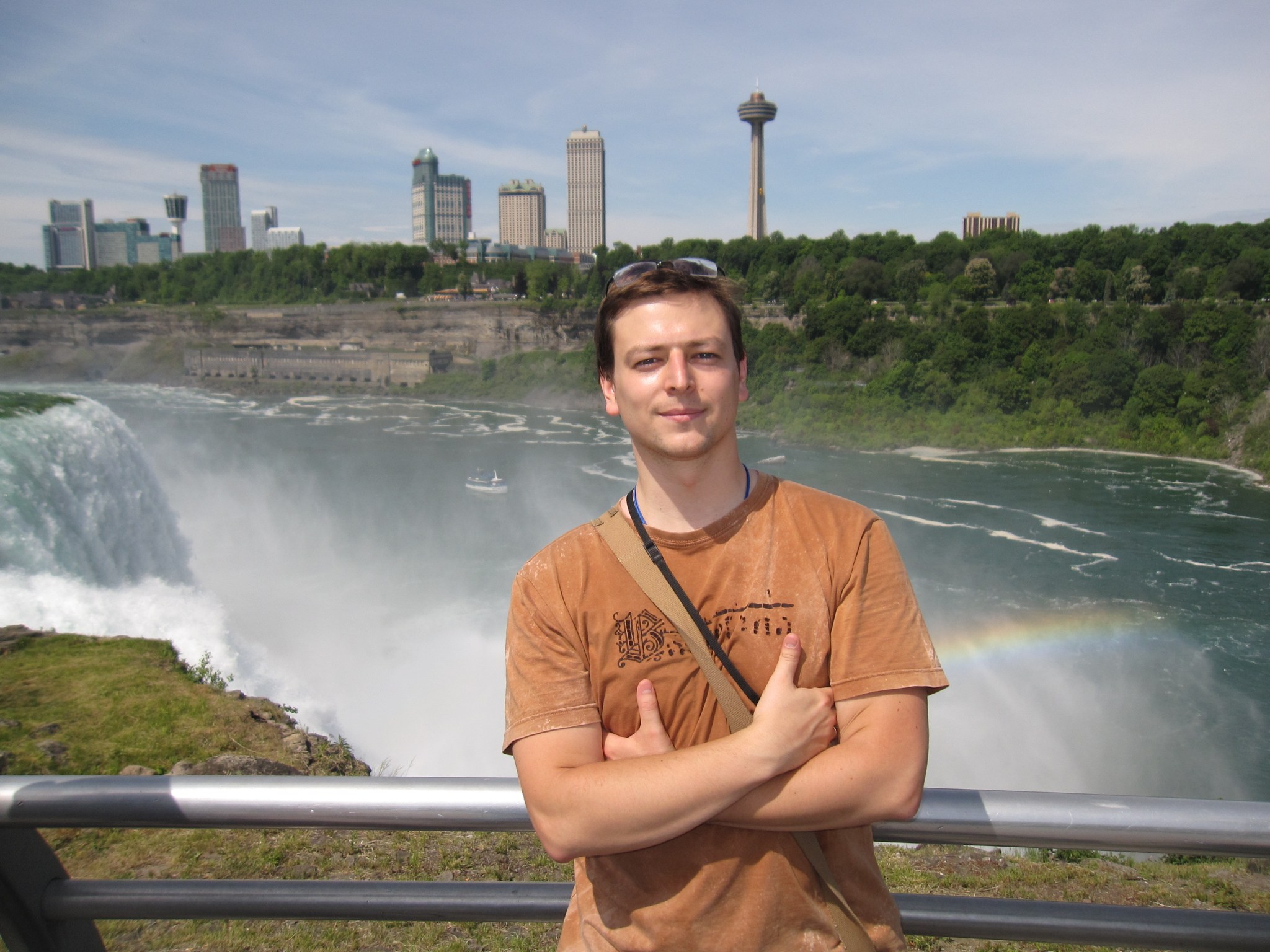Man with light skin and brown hair wearing a orange t shirt stands in front of a waterfall with skyscrapers along the horizon