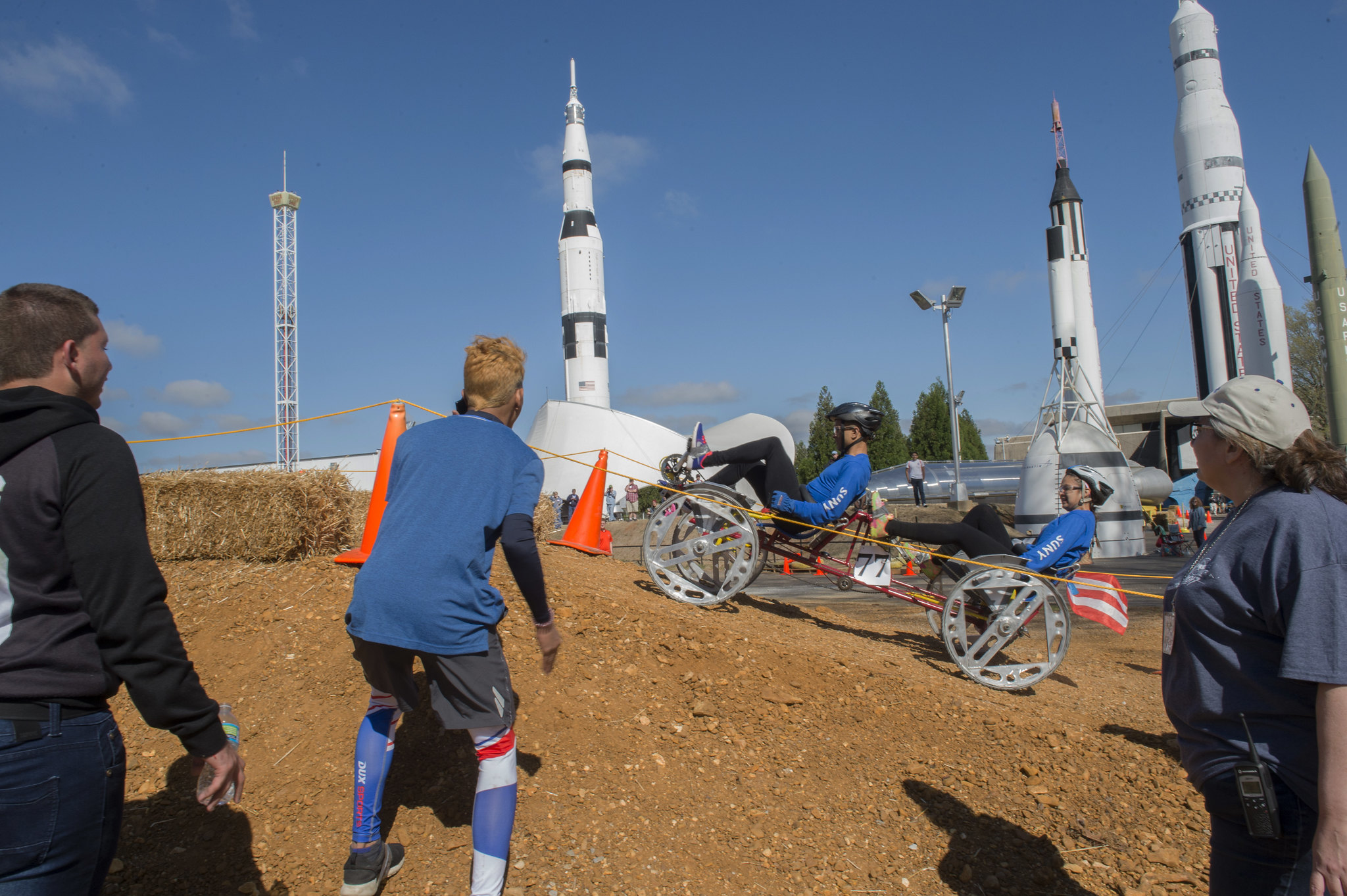 2 people on a human-powered rover with rockets in background