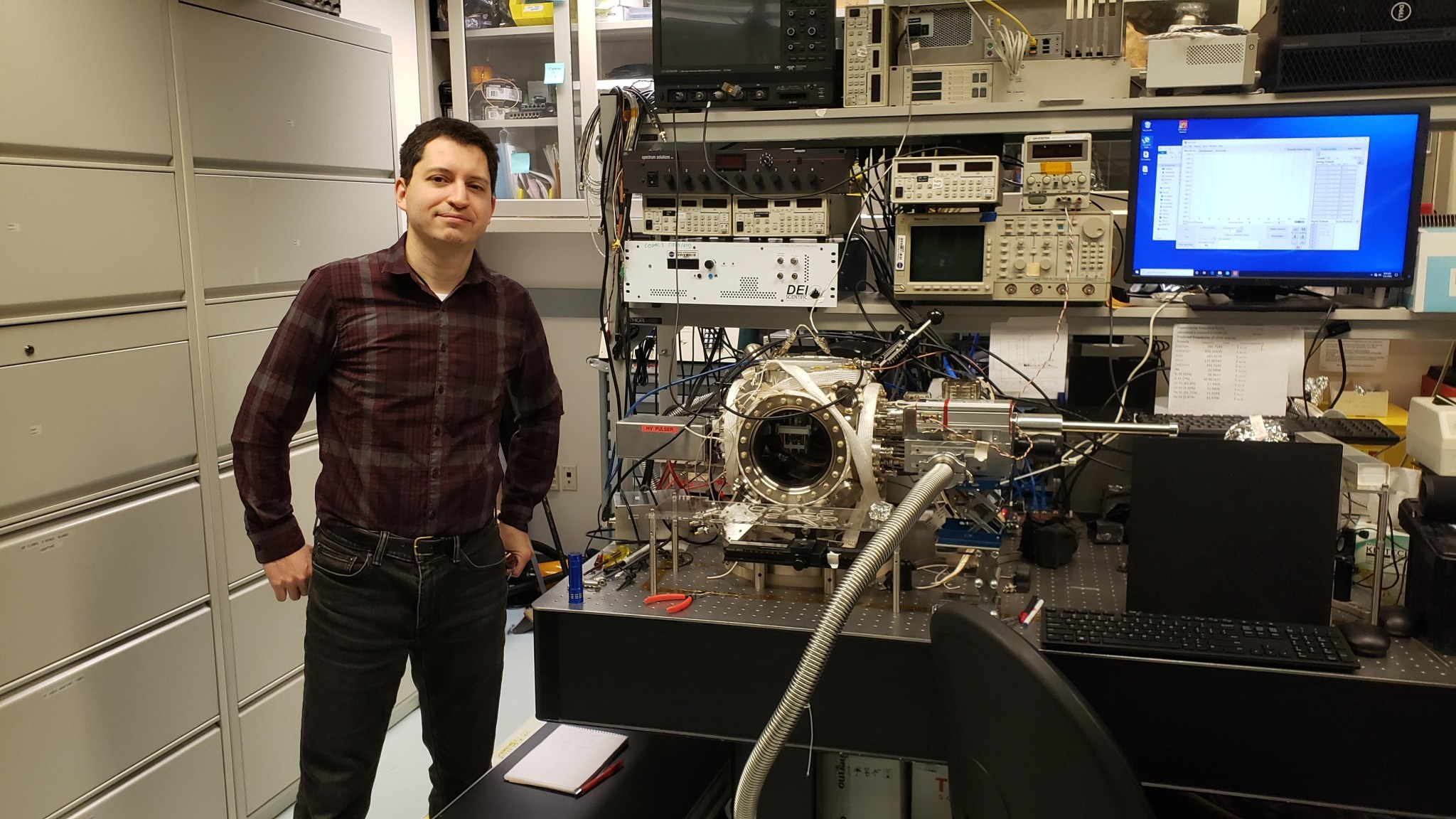 man with tan skin and black hair wearing a burgundy and black plaid shirt and jeans stands with his thumbs in his pockets, smiling at the camera. He is standing next to lab equipment and a keyboard with monitor.