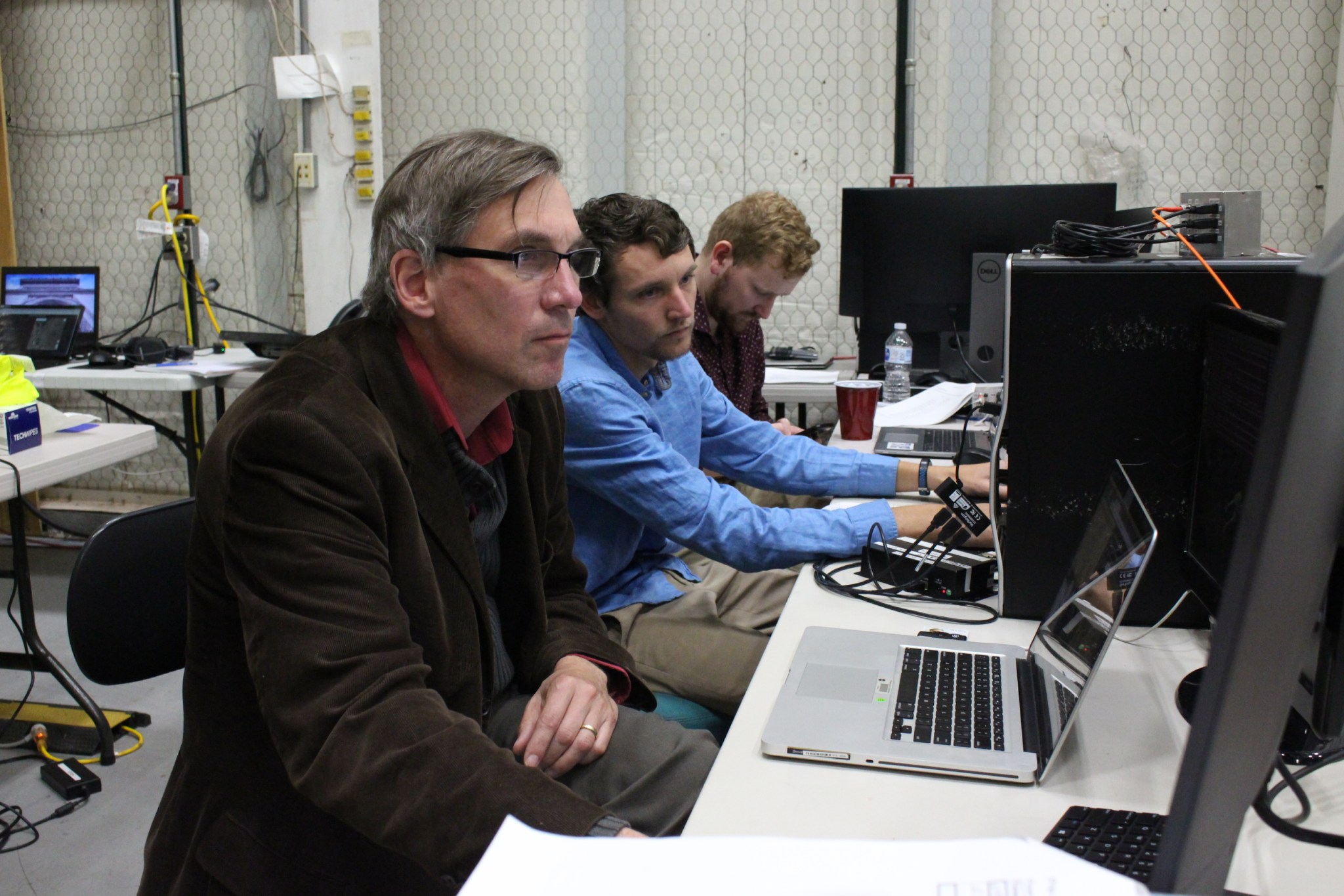 three men at a desk with many computers. The man closest to the camera is wearing a brown blazer and glasses, the man to his left is wearing a blue dress shirt, and the third man is mostly blocked by the man in the middle. The two men on the left are looking at the same screen. 