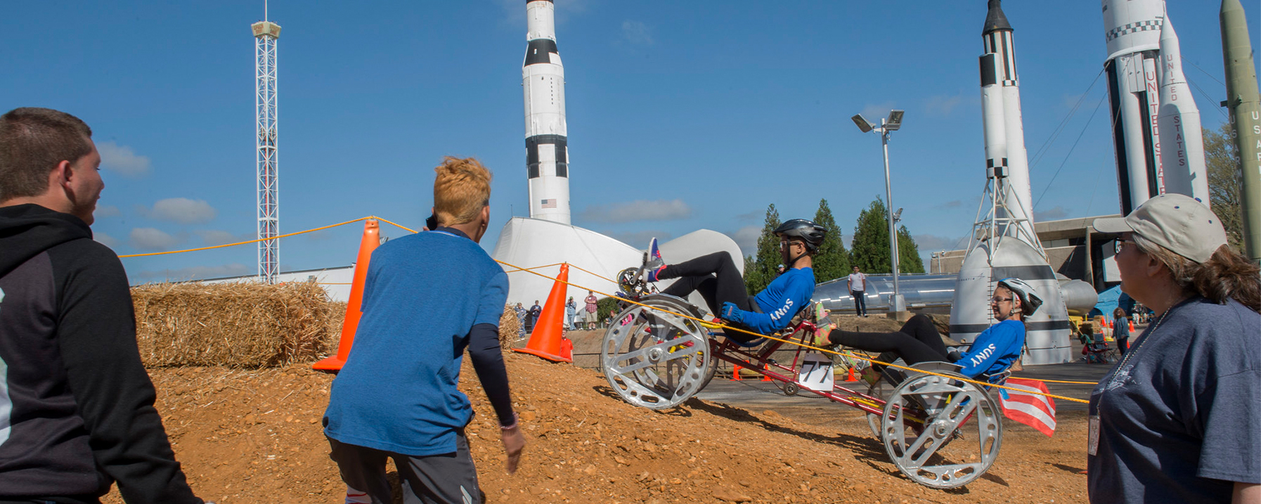 2 people on a human-powered rover with rockets in background