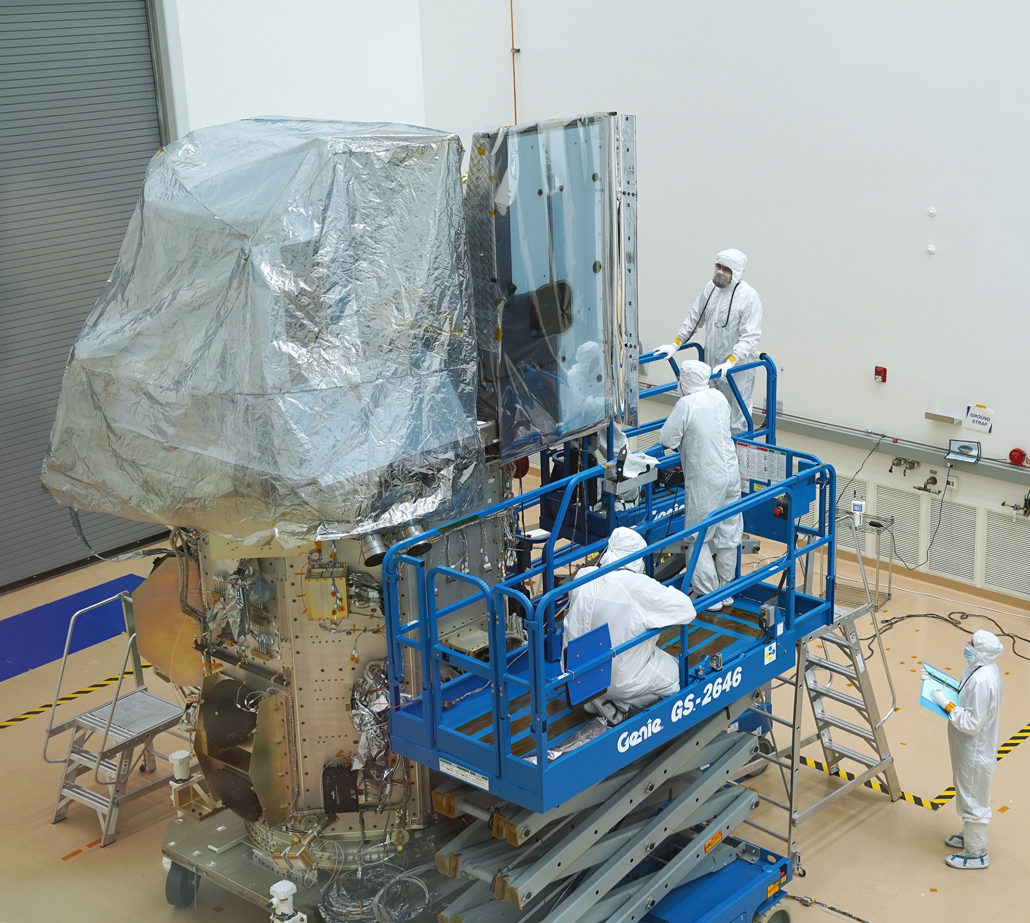 The Landsat 9 satellite stands in a cleanroom, with foil covering part of the top. The solar panel extends out one side, covered in clear plastic. Engineers in white cleanroom suits with masks and hoods stand and crouch on blue mechanical lifts, inspecting the satellite.