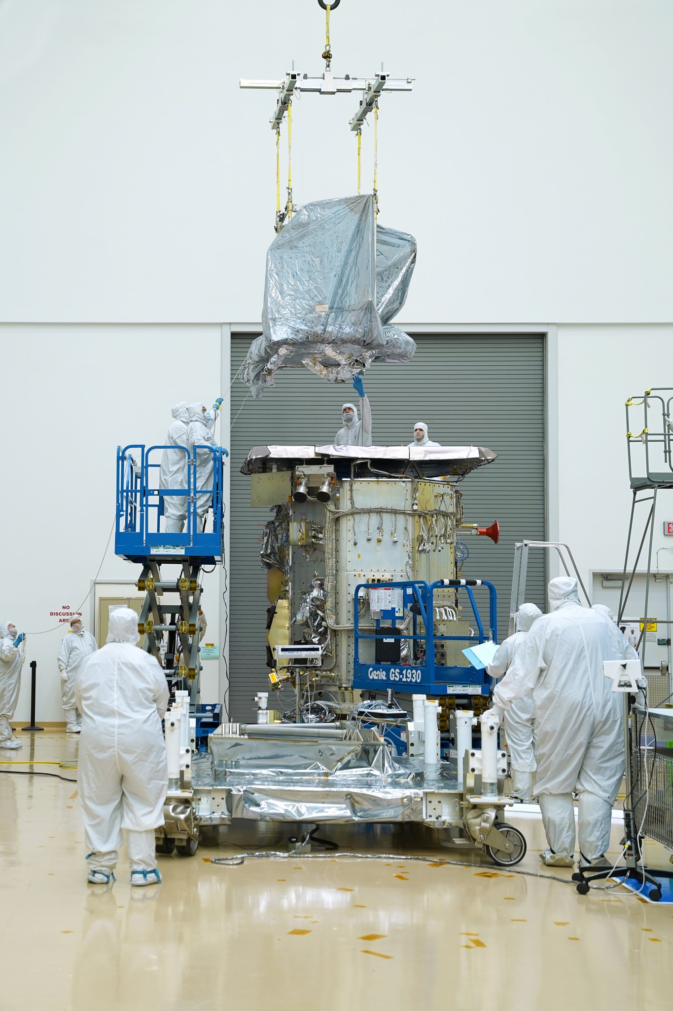 One of Landsat 9's instruments is lowered by straps toward the spacecraft body. The spacecraft is in a white clean room. Engineers in white cleanroom suits with hoods and masks are standing around the spacecraft body, as well as on blue mechanical lifts, helping lower the instrument.