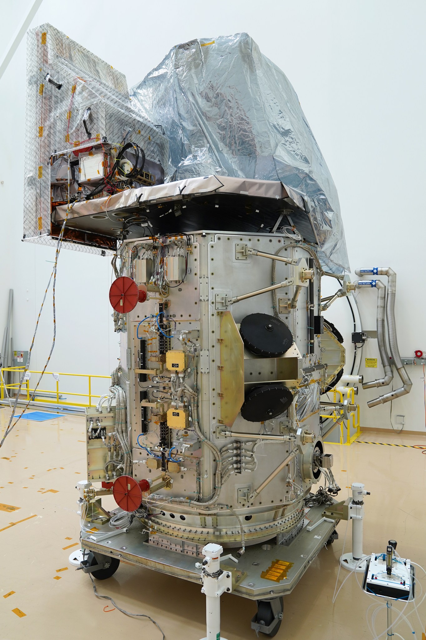 The Landsat 9 spacecraft sitting in a white clean room on a tan floor. The spacecraft is a silver cylinder with electrical components coming out all along the sides.