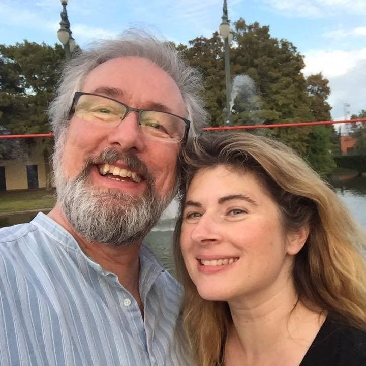 A man with fair skin and gray hair and a gray and black facial hair wears a blue striped shirt and glasses. He is smiling and leaning his head against a woman with light skin and blonde/brown hair wearing a black shirt. They are both smiling. 