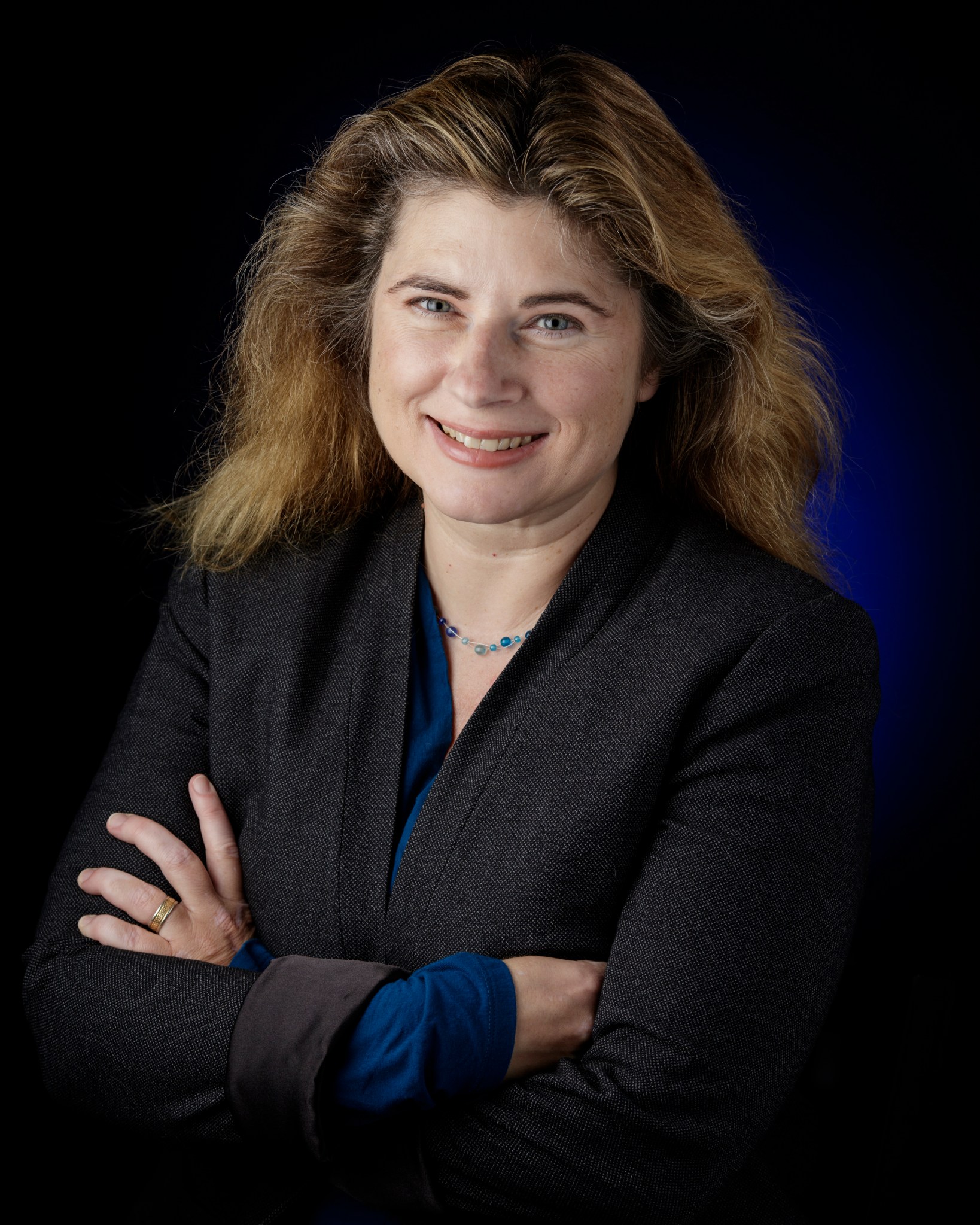 Woman with fair skin and long brown/blonde hair wears a blue shirt and black textured blazer, along with a gold wedding ring. She is smiling and standing against a black and blue background. 