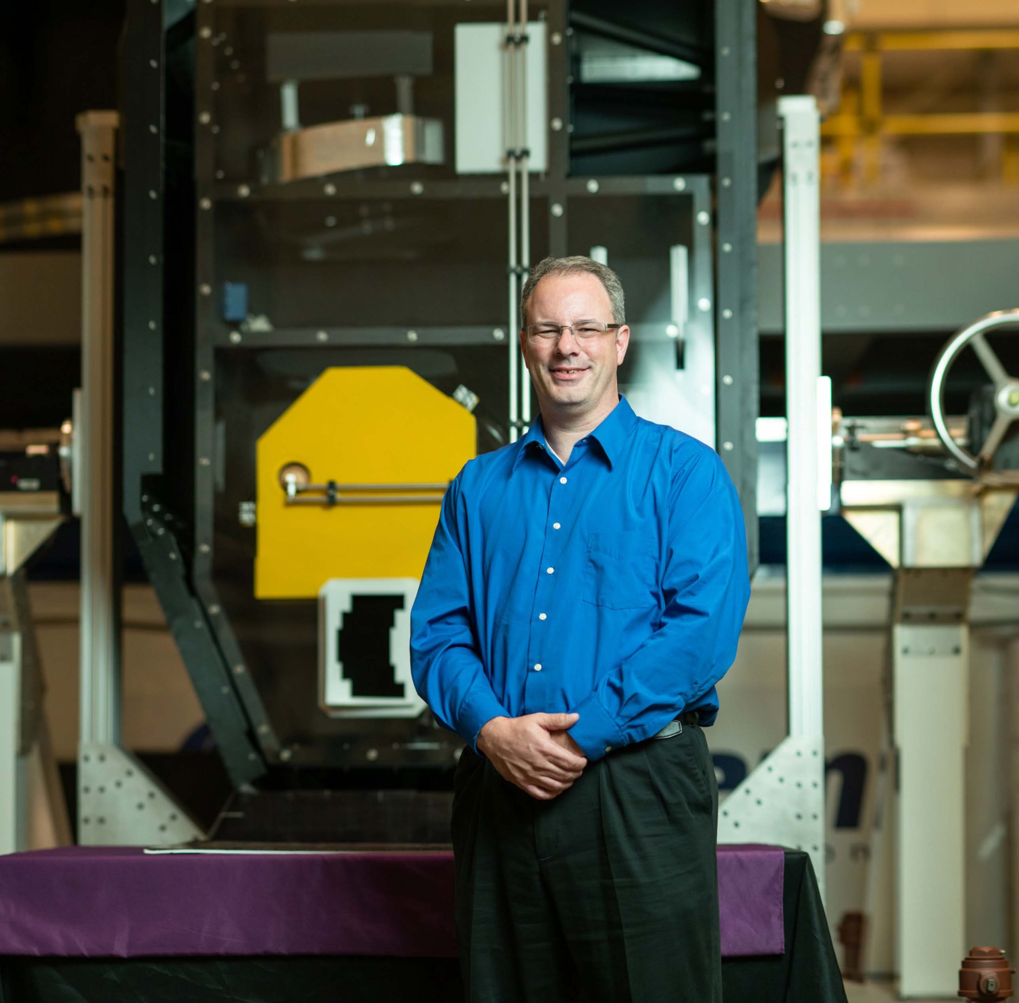 Man with shirt grey-brown hair and glasses, wearing a blue dress shirt and black pants stands, smiling, in front of machinery with one of the Webb, gold hexagonal mirrors inside.  