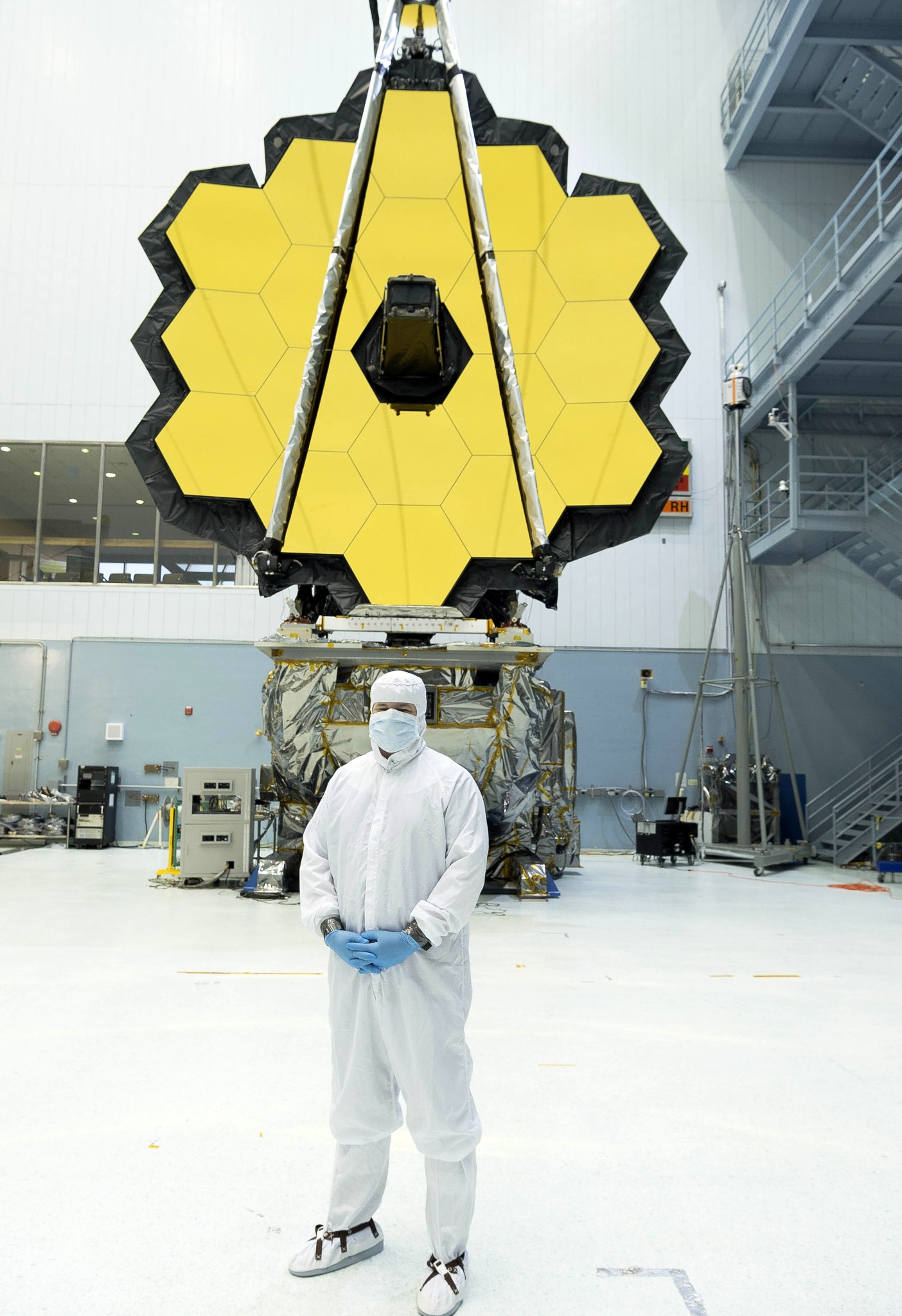 Jason Hylan, dressed in an all white, sterile "bunny suit," stands in front of the Webb telescope in a cleanroom. 