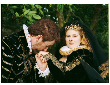 A man with fair skin and brown hair wears formal Renaissance clothing and is kissing the hand of a woman with hair skin and blonde hair, wearing formal Renaissance clothing and a crown, she softly smiles at the camera. They are standing in front of lush green leaves on trees. 