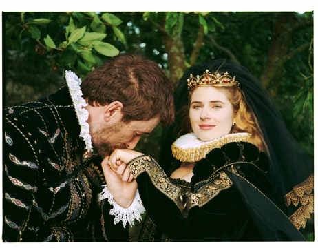 A man with fair skin and brown hair wears formal Renaissance clothing and is kissing the hand of a woman with hair skin and blonde hair, wearing formal Renaissance clothing and a crown, she softly smiles at the camera. They are standing in front of lush green leaves on trees.