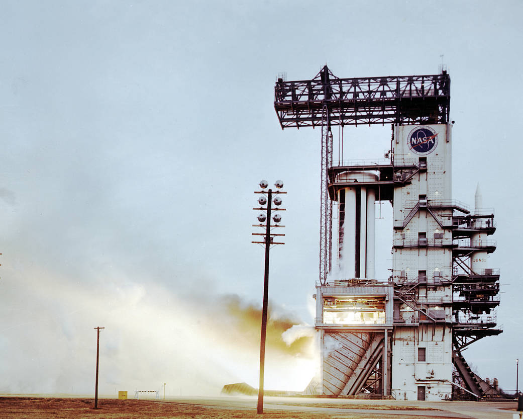 Saturn I rocket test stand firing. The image has a large test stand with flames pointing to the left of the image.