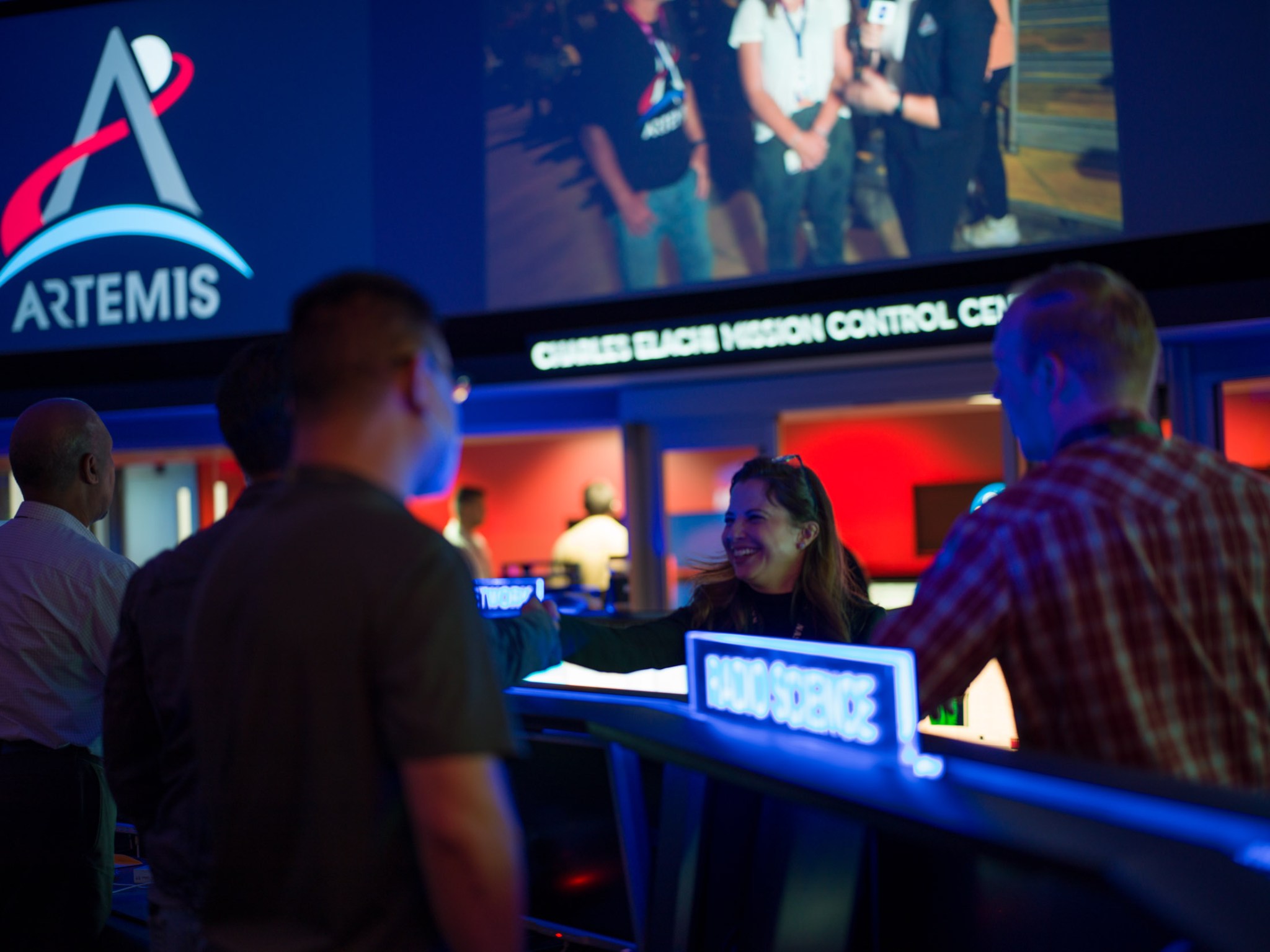 Five team members stand talking in the Charles Elachi Mission Control Center at NASA's Jet Propulsion Laboratory. A sign in the foreground reads "Radio Science." The Artemis logo can be seen on a large screen in the background.