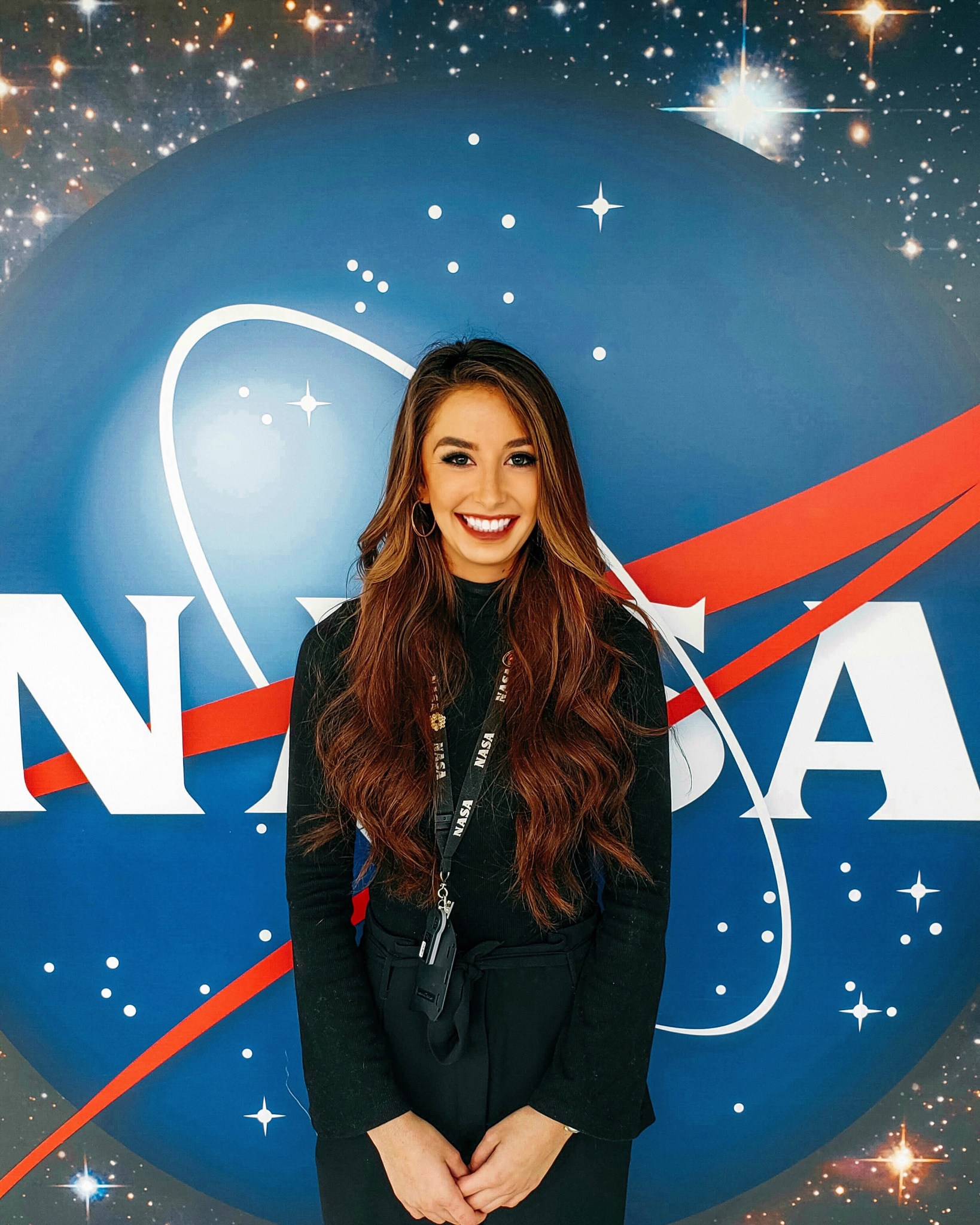 Woman with long, dark, wavy brown hair wearing a black long sleeve shirt, black pants, and a black NASA lanyard stands, smiling, in front of a large NASA meatball sign.