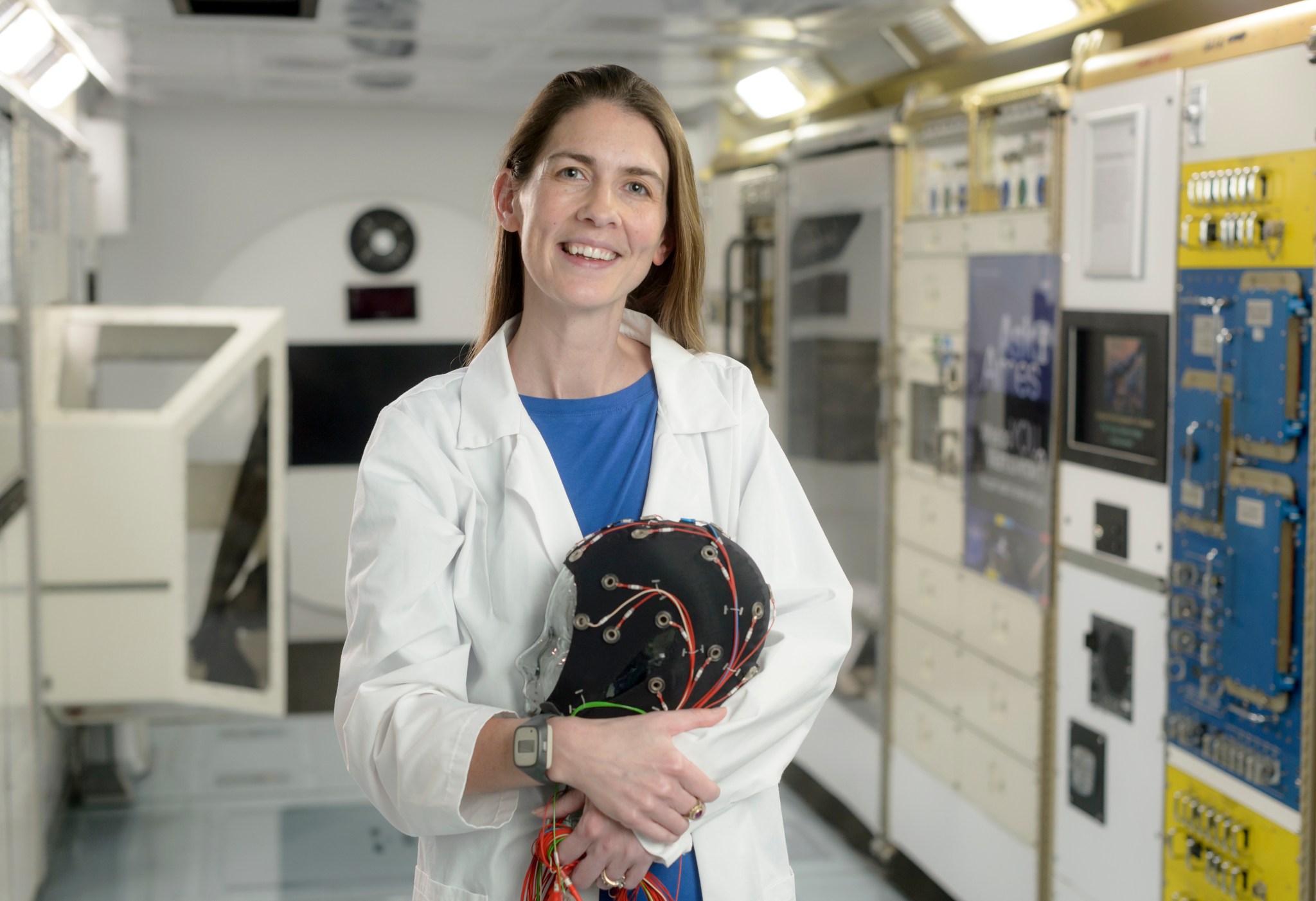 A woman in a white labcoat stands in a lab holding a cap covered in electrodes