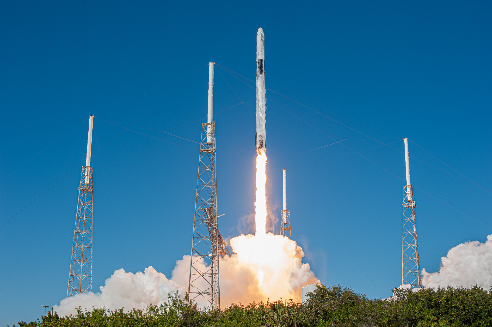  A Falcon 9 rocket launching against a flat, bright blue sky. The rocket is a thin white cylinder with black stripes. Bright yellow white flames erupt out the bottom of the rocket, disappearing into a cloud of white steam. Four towers encircle the launchpad.