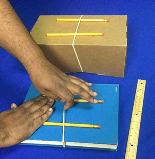 Hands pluck a rubber band that is wrapped around a book and two pencils that are below a ruler on the table