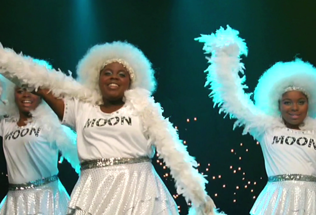 Three girls dancing and wearing white outfits with the word moon on their shirts