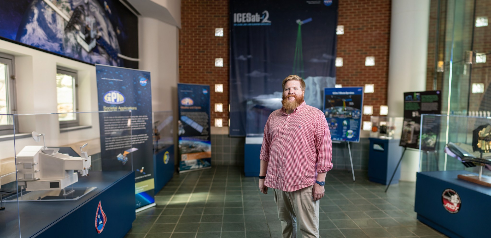 Man with auburn hair on his head and face, wearing a red shirt and tan pants stands with models of satellites and in front of a sign that says ICESat-2