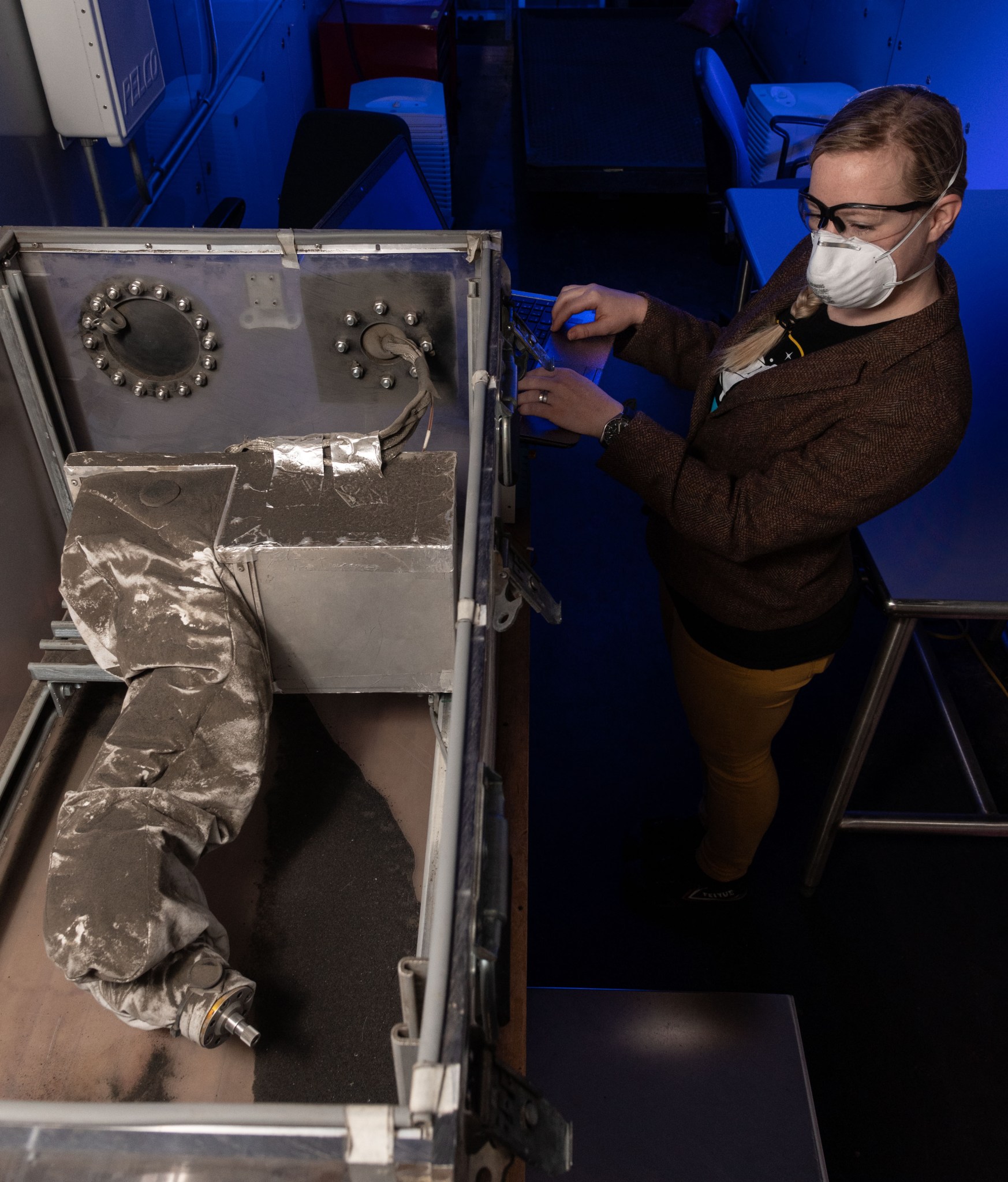 A  woman wearing a dust mask stands next to a test chamber containing simulated lunar soil and rover wheel module components