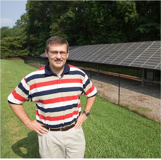 mans stands in grass with solar panels to his left. He's wearing a navy, tan, red, and white striped polo.