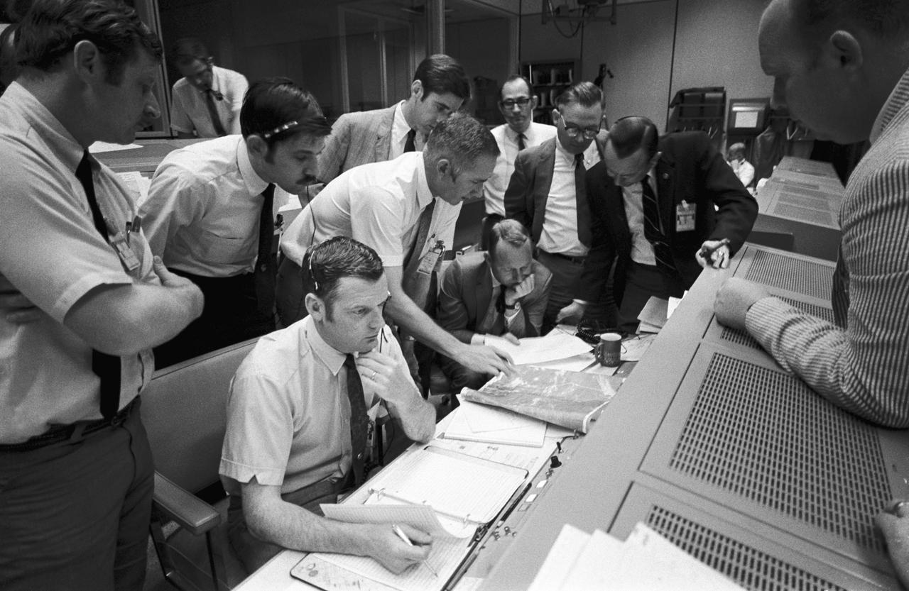 A group of flight controllers gathers around the console of Glenn S. Lunney (seated, nearest camera)