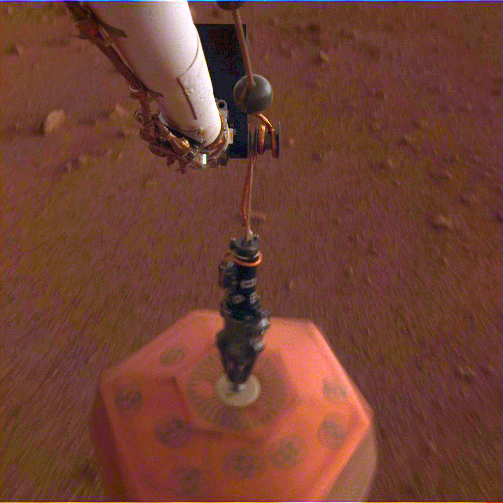 The seismometer on the MarsInsight mission. The ground is red dirt and the instrument is a large orange hexagonal object. 