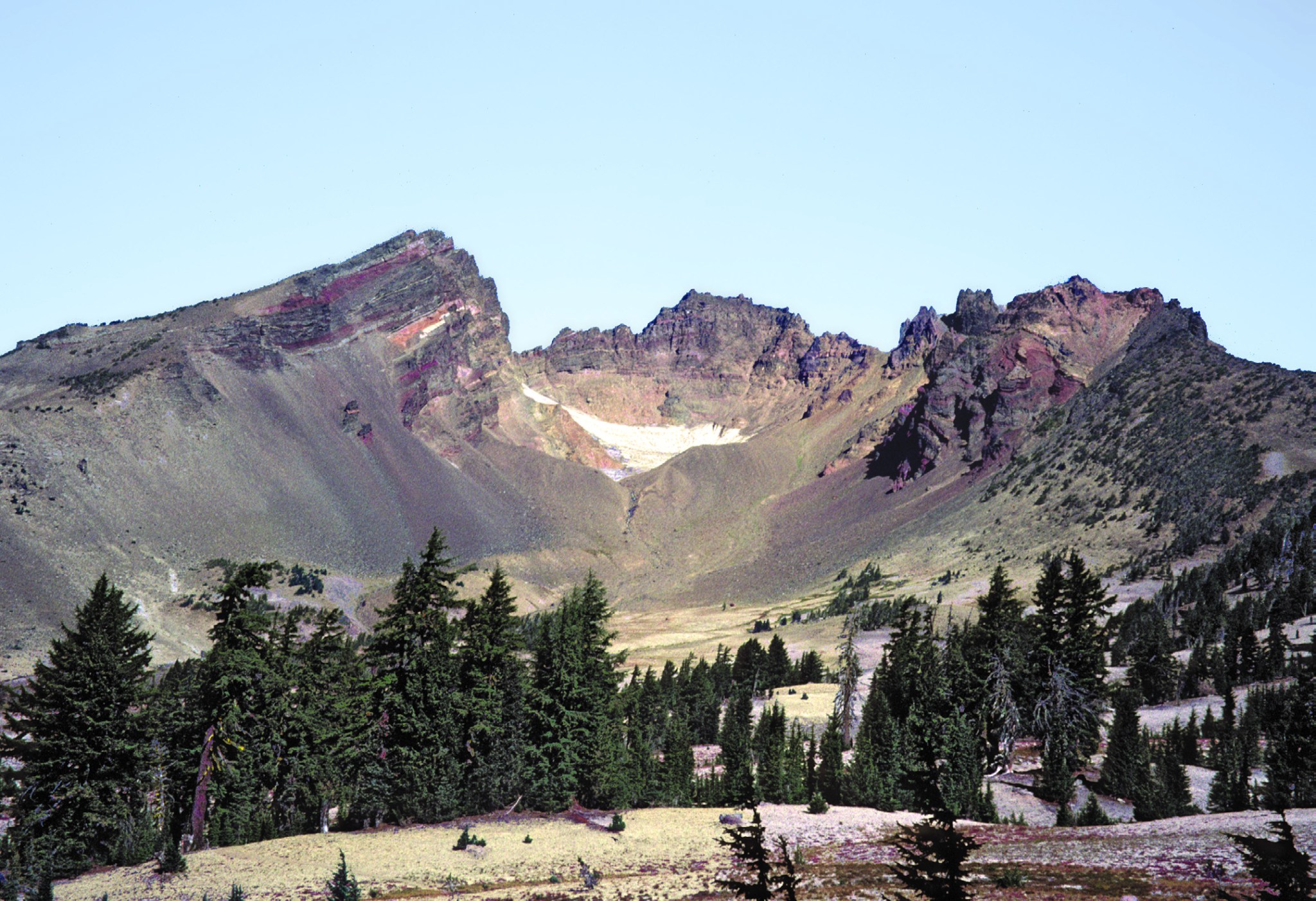 Three Sisters Volcano in Central Oregon. The mountain is a dark rainbow of colors, with purples and browns fading into pinks and greens. In the caldera, white peeks out. A line of pine trees stands in the foreground. 