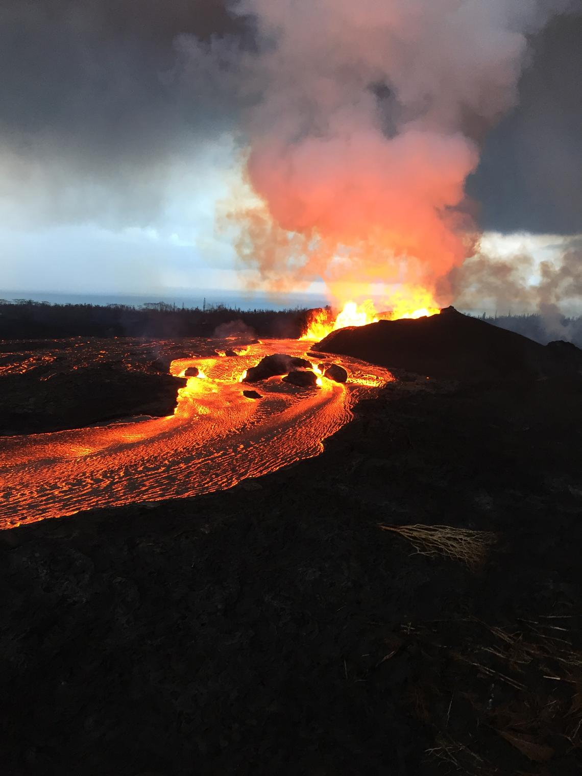 photo of orange lava flwoing over the ground from Kilauea vent in the distance