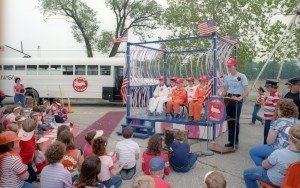 Teacher stands next to podium with students in space flight suits with crowd in foreground.