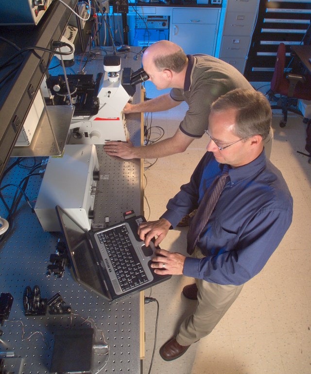 Two researchers, one at a computer and the other at a microscope, in a NASA lab.
