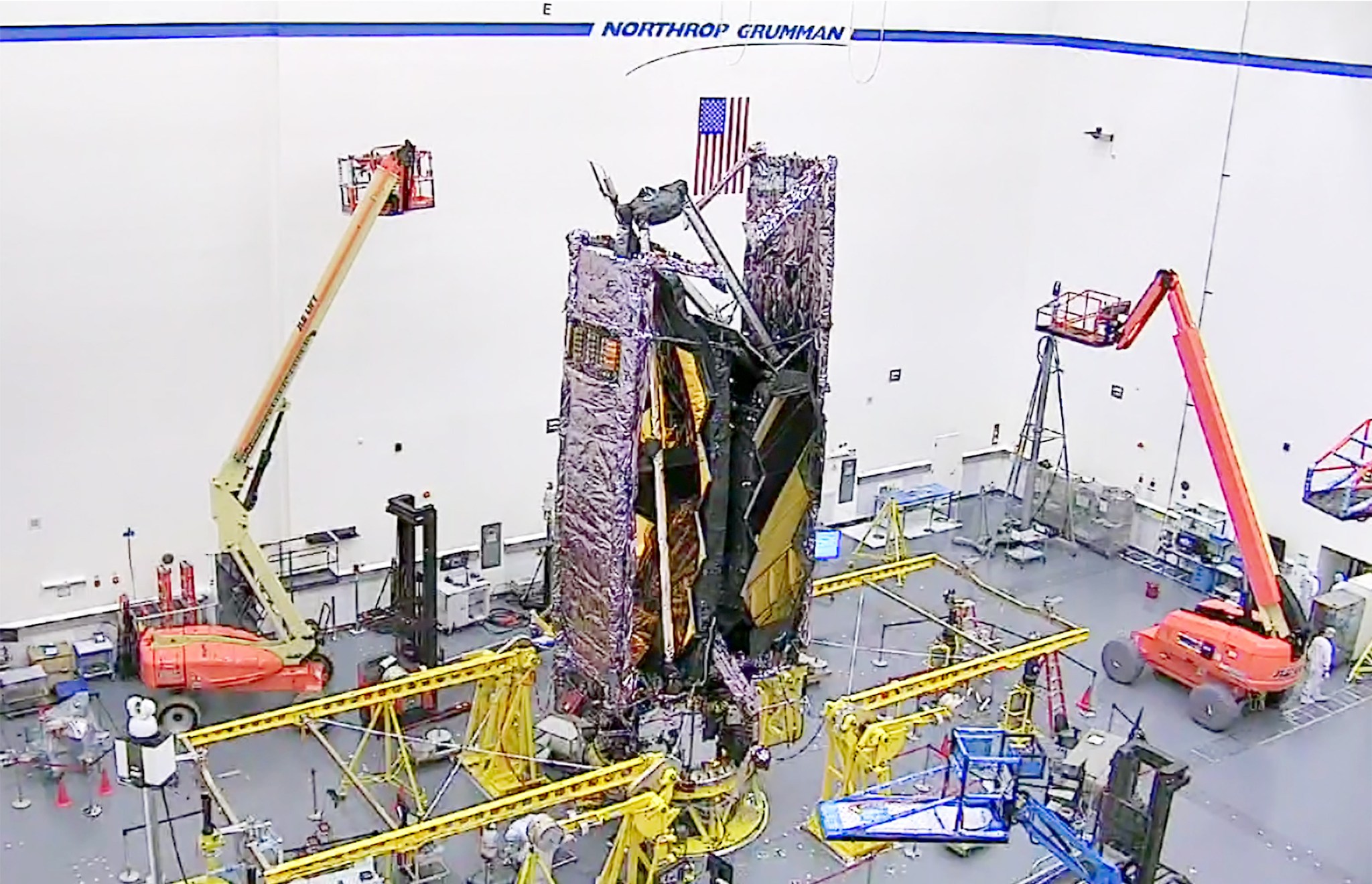 The James Webb Space Telescope in a cleanroom surrounded by multiple technicians and two boon lifts.