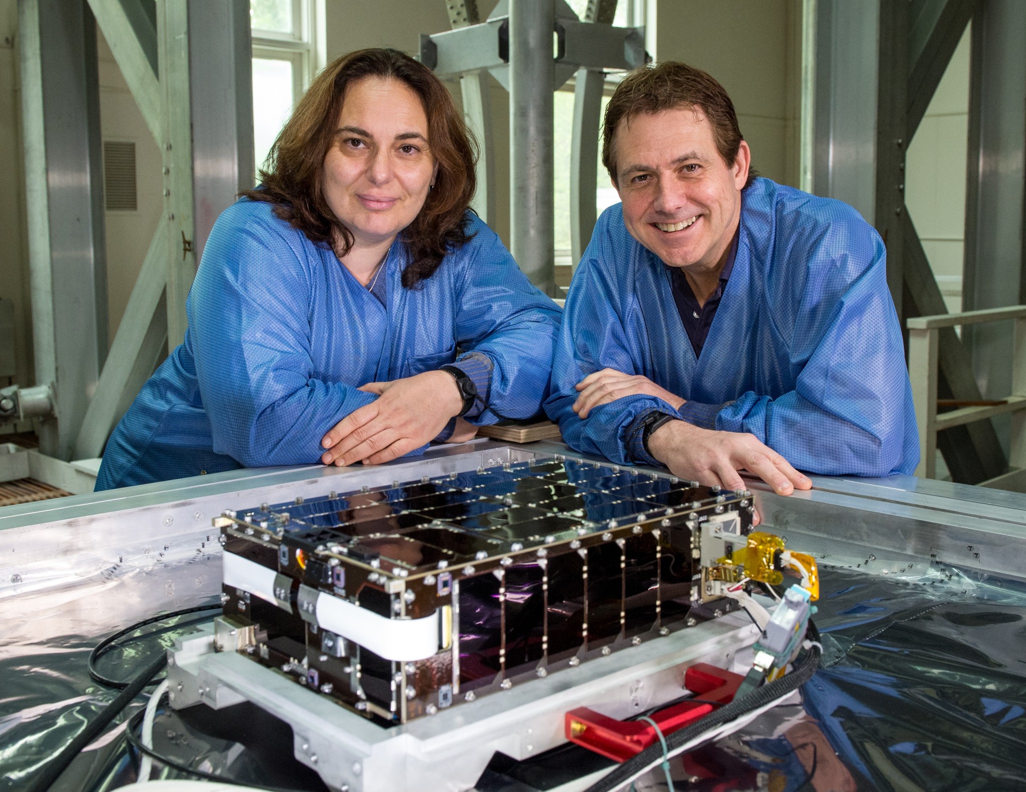 A woman on the right and a man on the left wearing blue lab coasts, smiling next to technology that will be used in the mission. 