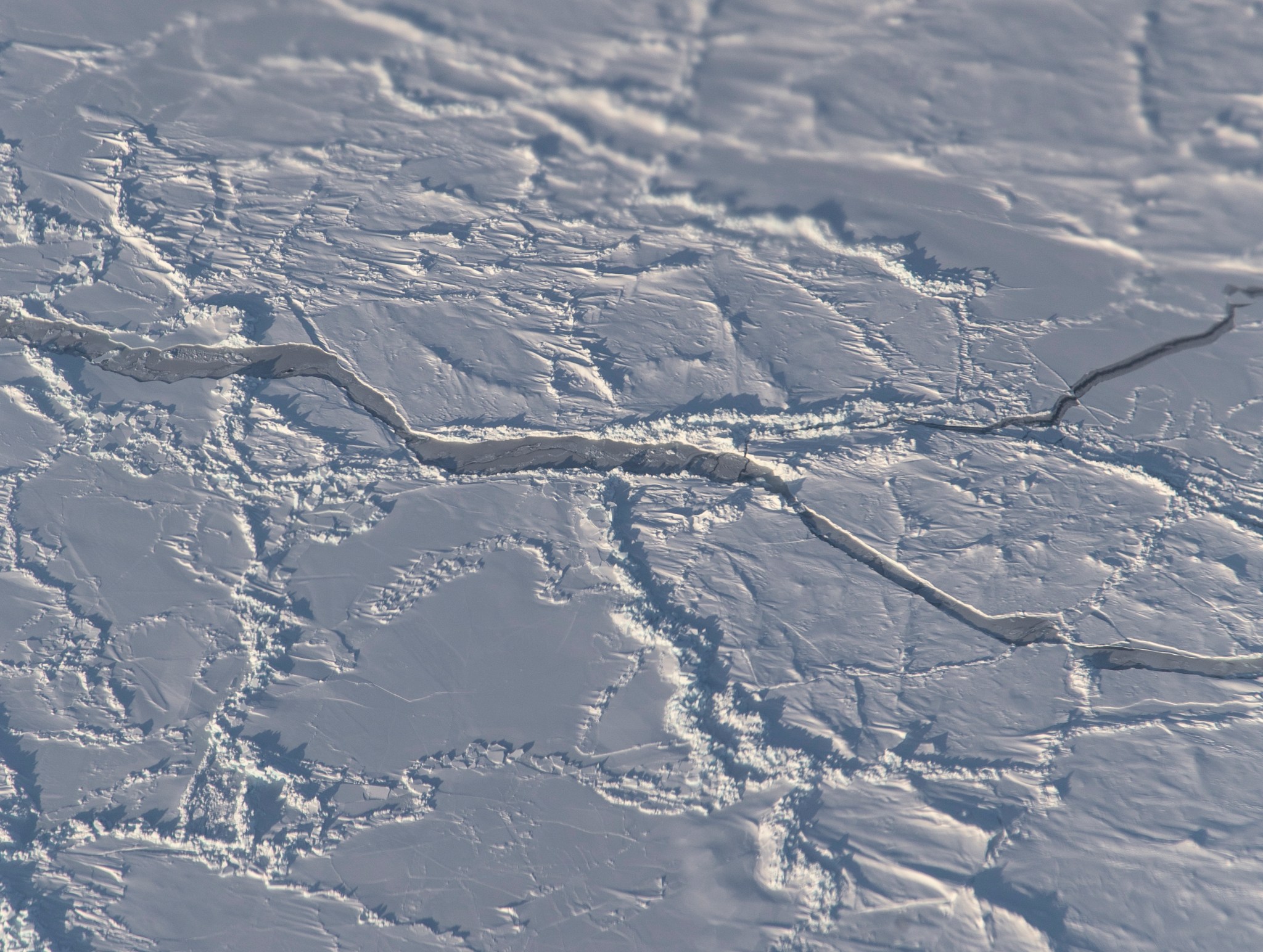 Arctic sea ice, with ridges and cracks. The ice is blueish white and mostly flat, with jagged cracks and ridges running across it.