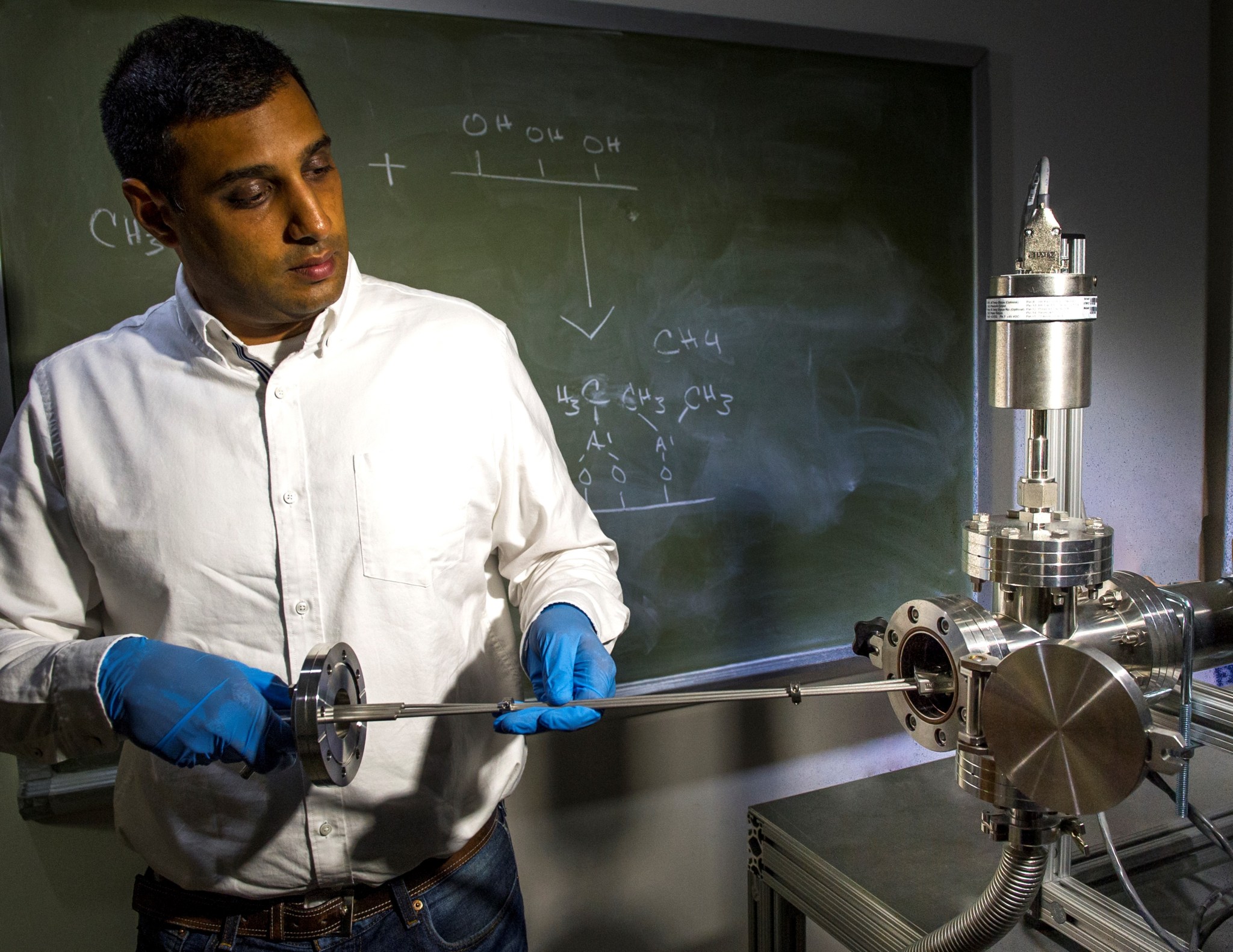 Man with dark tan skin and short black hair holds a metal rod attached to a machine. He is wearing  a white button down shirt, jeans, and blue gloves. There's a green chalk board with numbers and letters behind him.