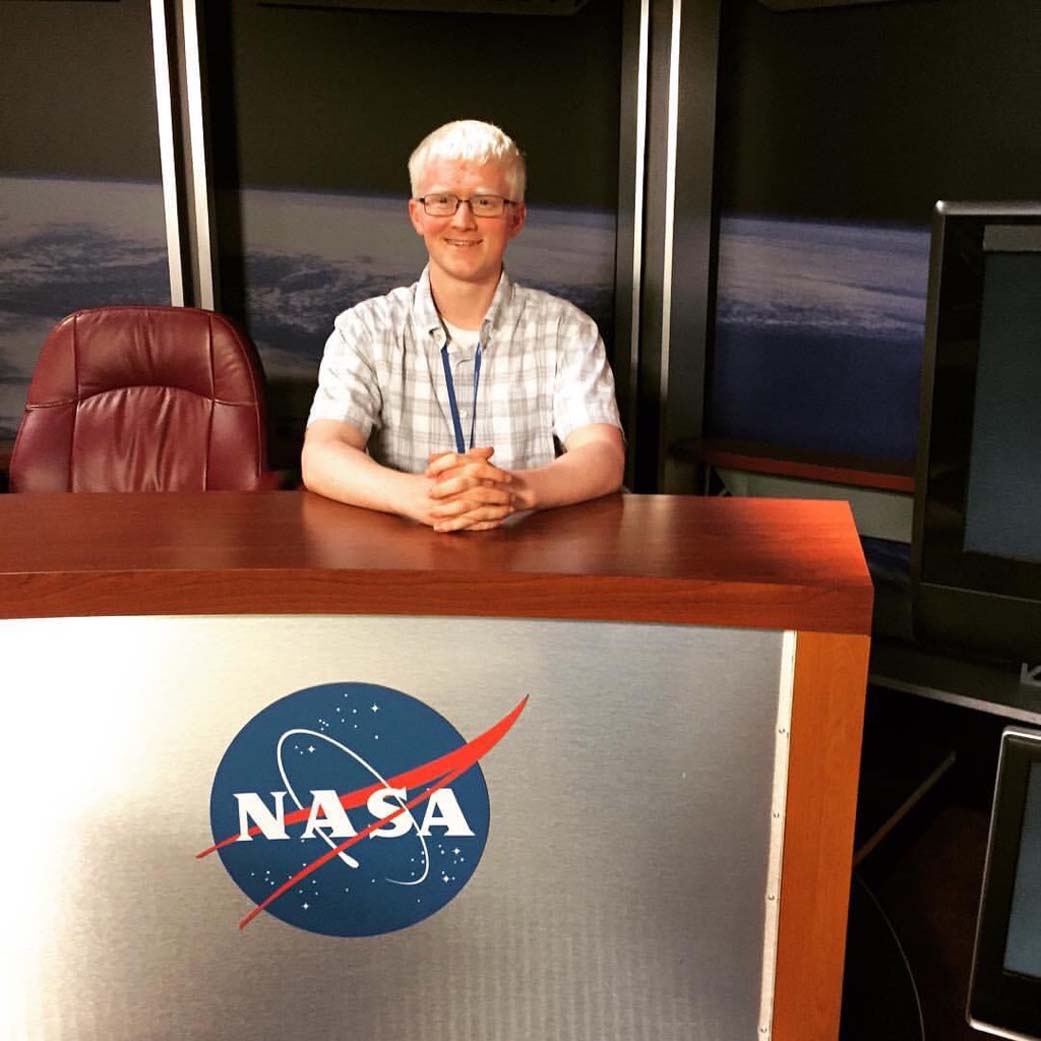 Man with fair skin and light blonde hair wears glasses and a short sleeve white plaid shirt sits  with his hands folded together at a desk with a large NASA meatball on the front and an image of Earth from space in the background. 