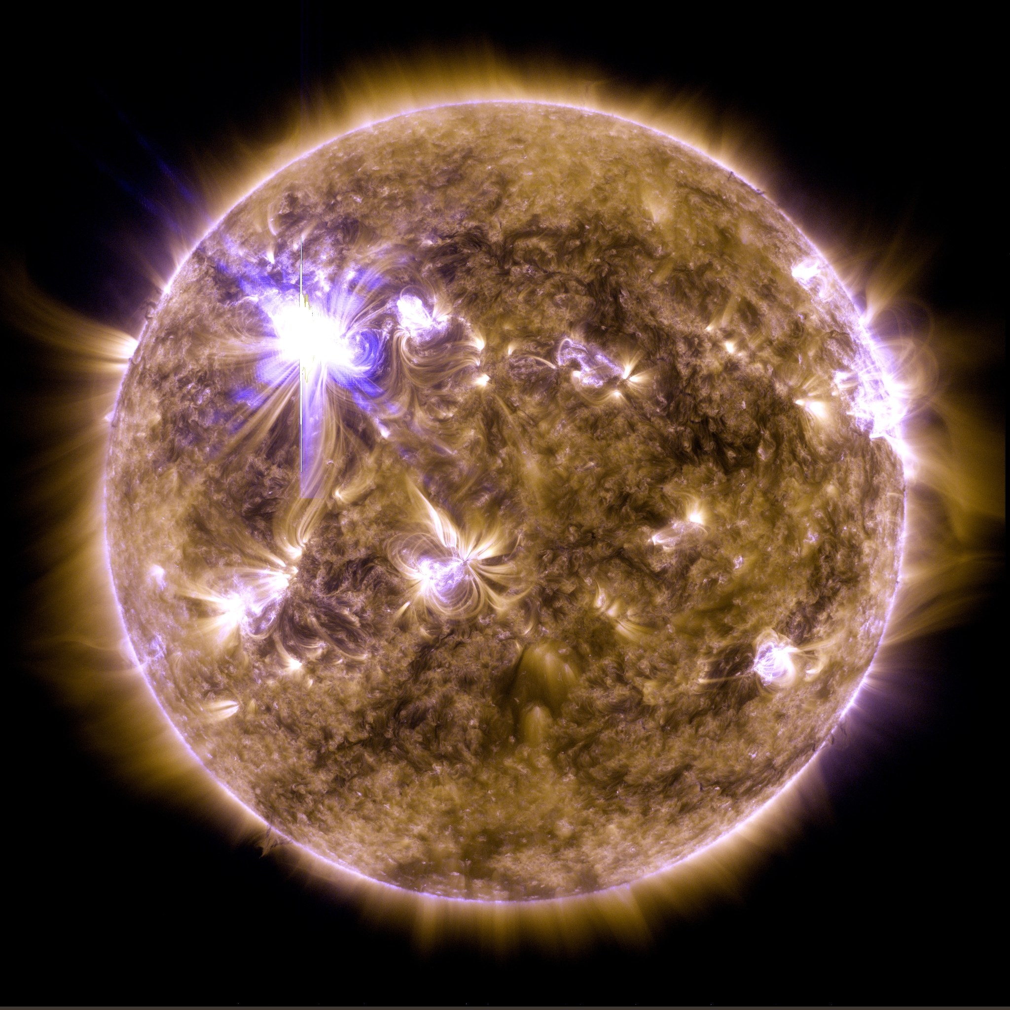 A satellite image shows the Sun colorized in gold and purple. A bright white burst of light shines in the upper left part of the Sun.