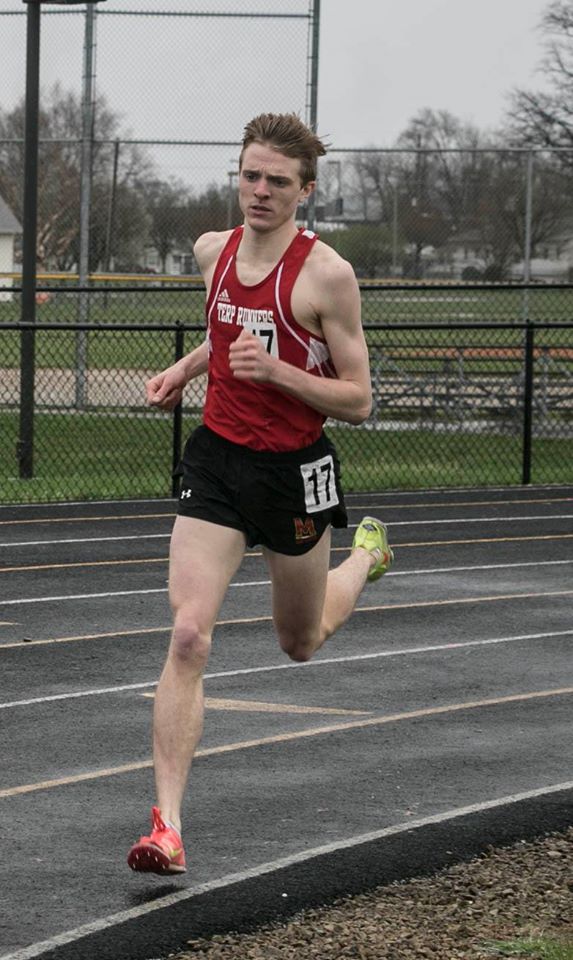 Man with fair skin and short brown hair wears a red tank top, black shorts, one orange racing spike shoe and one yellow spike shoe, runs on a track. He wears the number 17.  The photo is taken mid-stride and the runner has a very focused look on his face. 